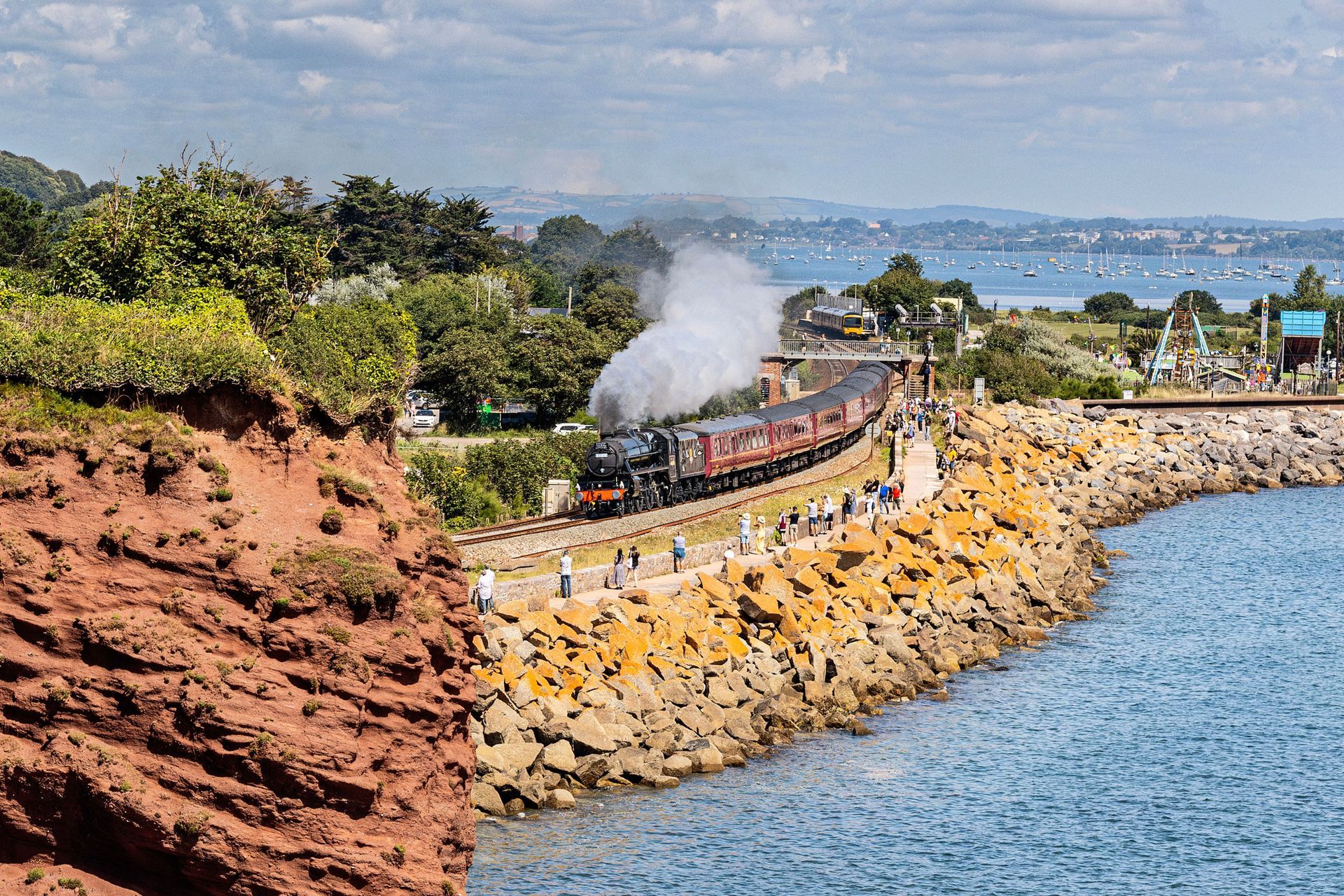 Steam train traveling along a seaside track; red rock cliff, ocean, and bay in the background.