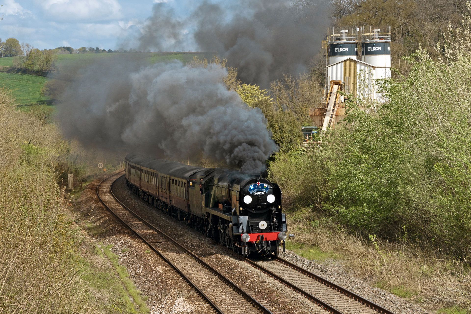 Steam locomotive pulling coaches through countryside, trailing thick black smoke beside tracks and trees