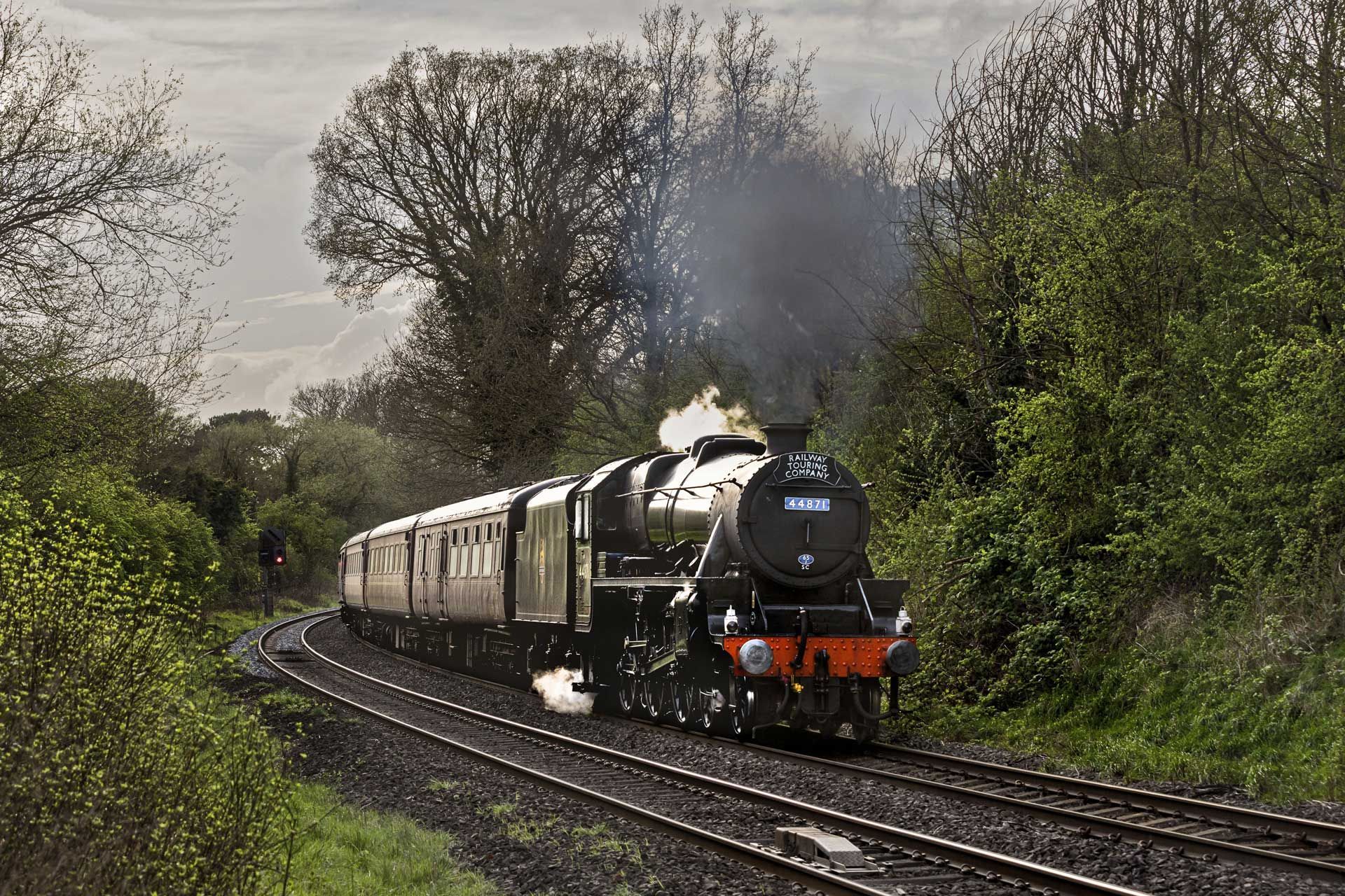 Steam train on tracks curves through a green, wooded landscape, puffing black smoke.