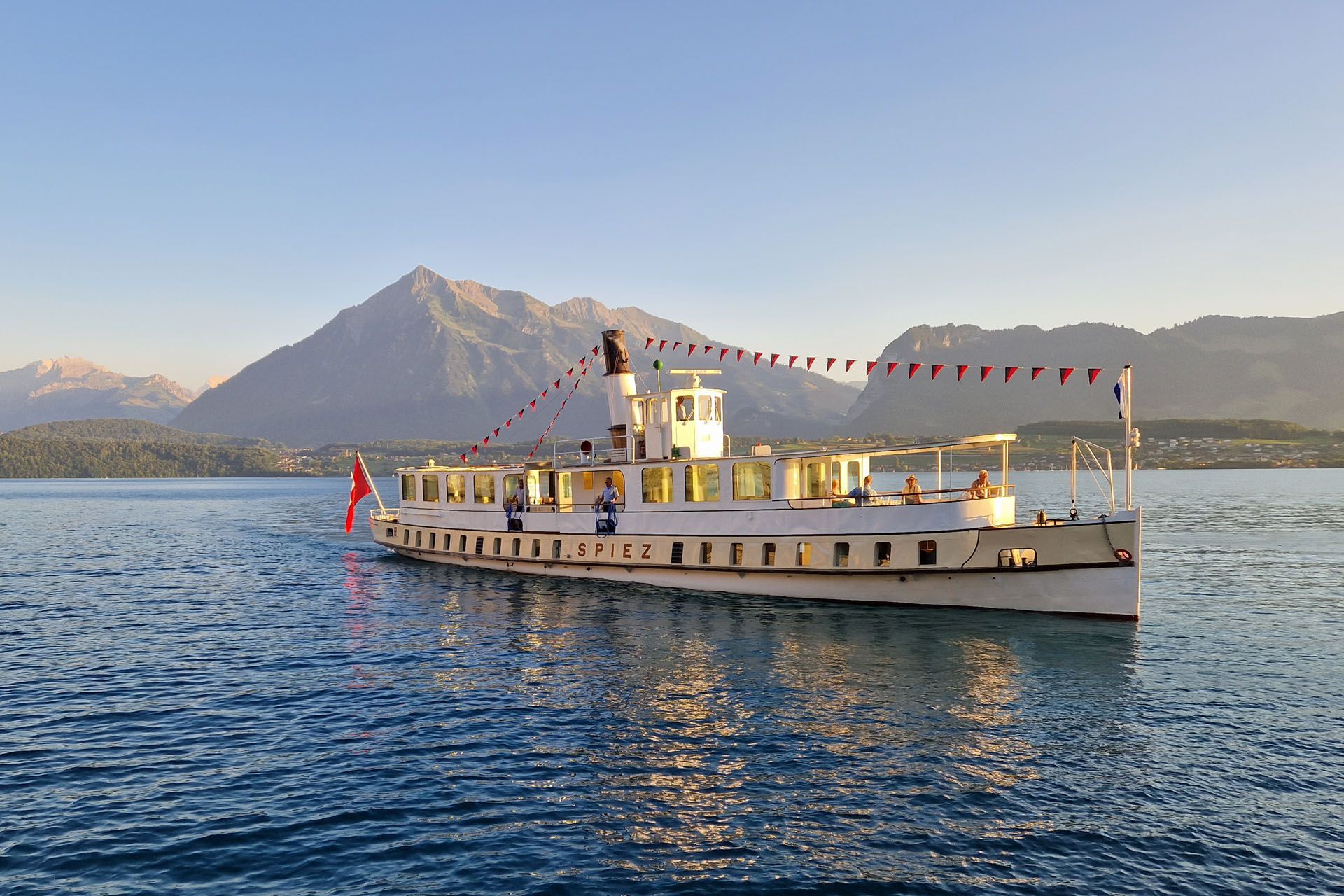 White boat on blue water with red flags, mountains and a hazy shoreline in the background.