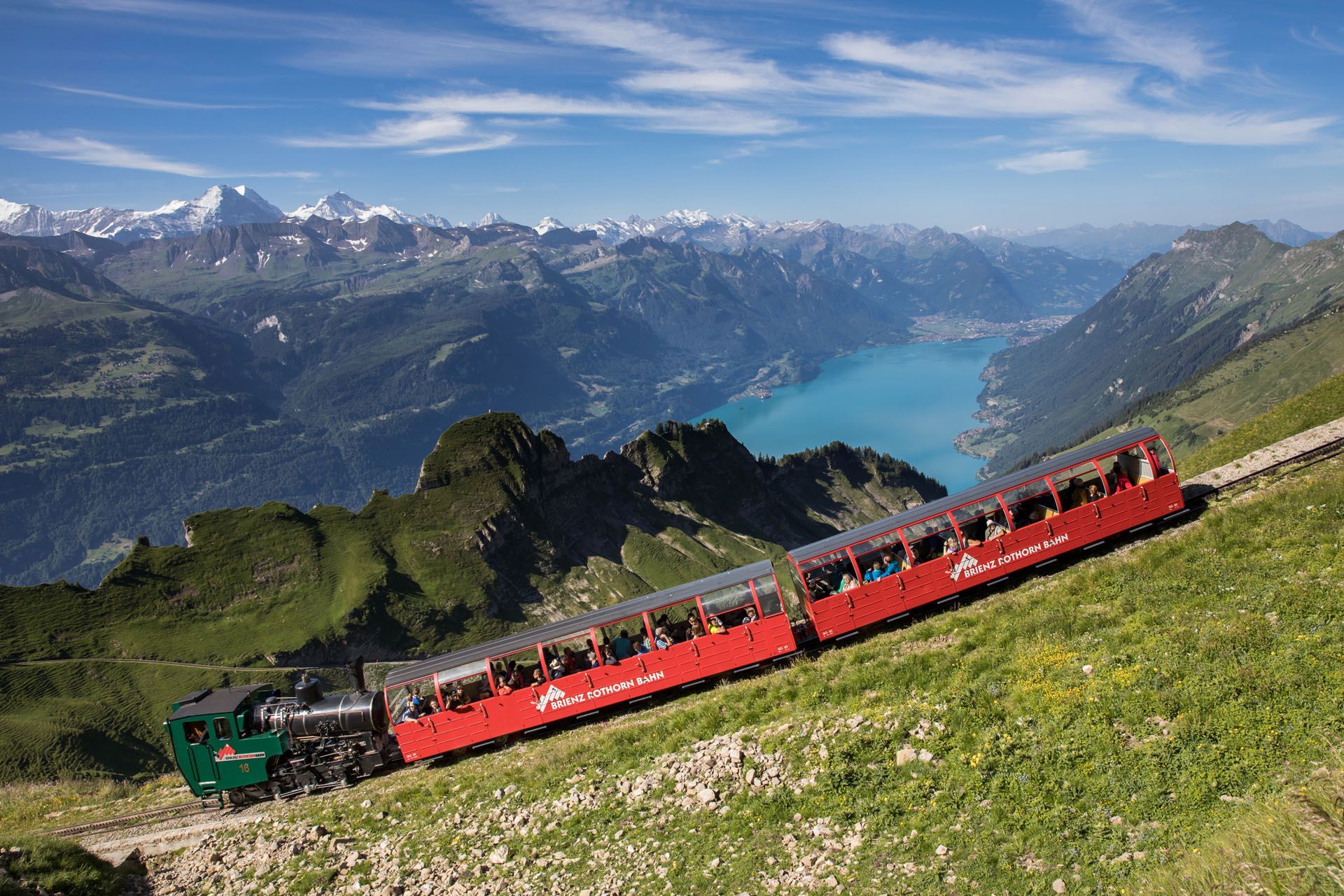 Red mountain train climbing a grassy slope with a turquoise lake and snow-capped peaks in the background