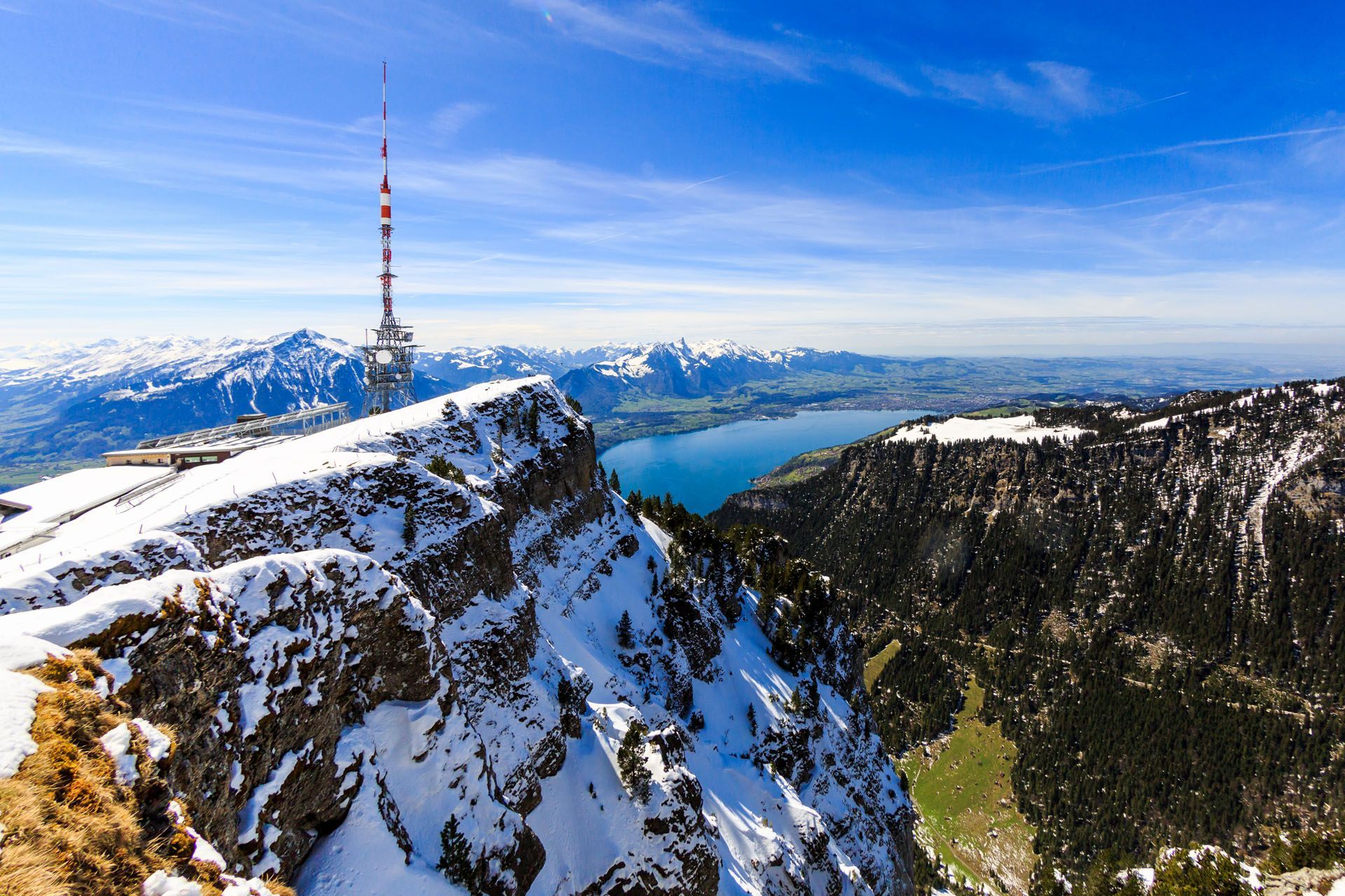 Snowy mountain ridge with a tall radio tower overlooking a blue lake and distant peaks