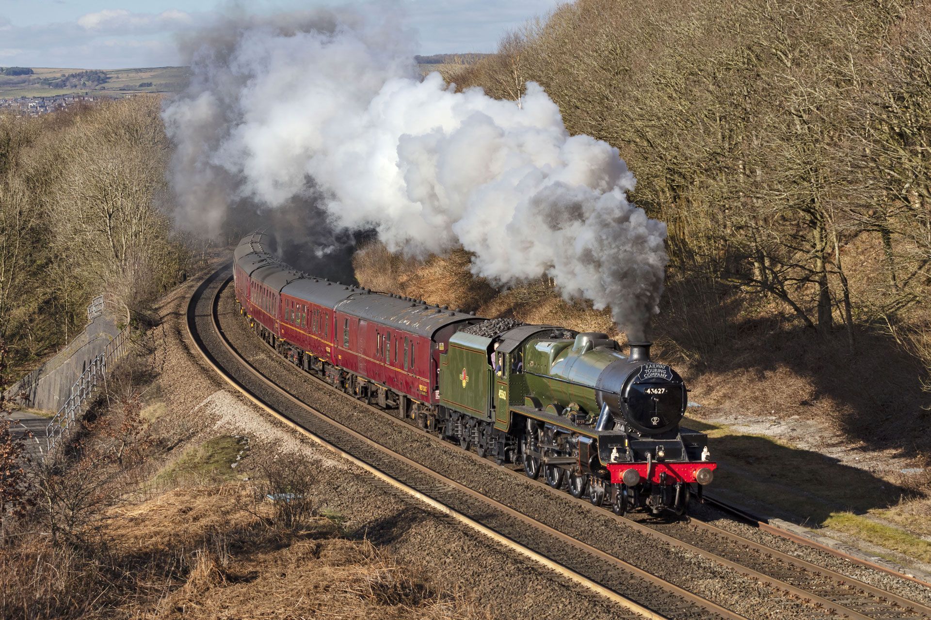 Steam train pulling red carriages through a countryside curve with billowing white smoke