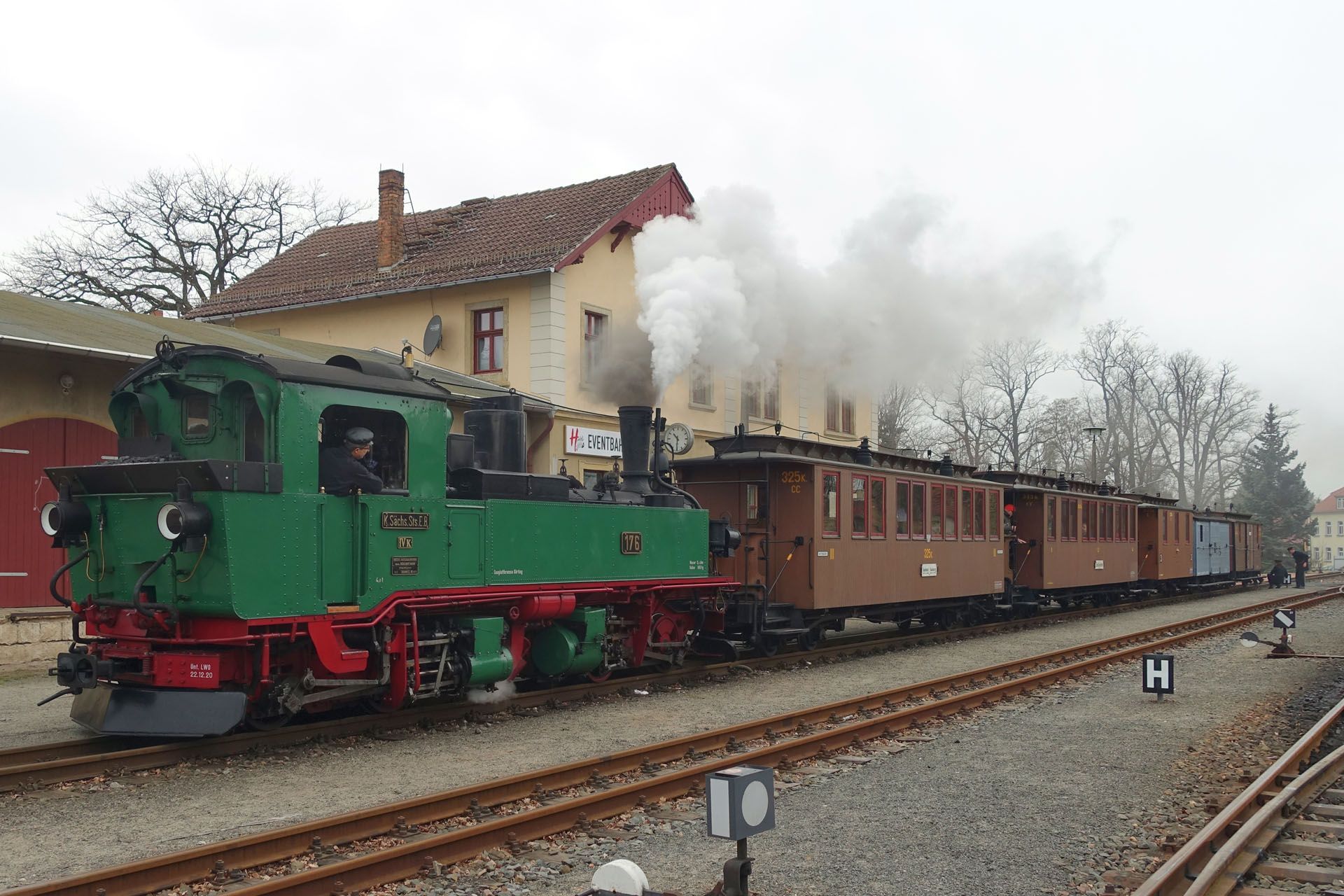 Green steam locomotive pulling brown passenger cars past a station on rainy tracks