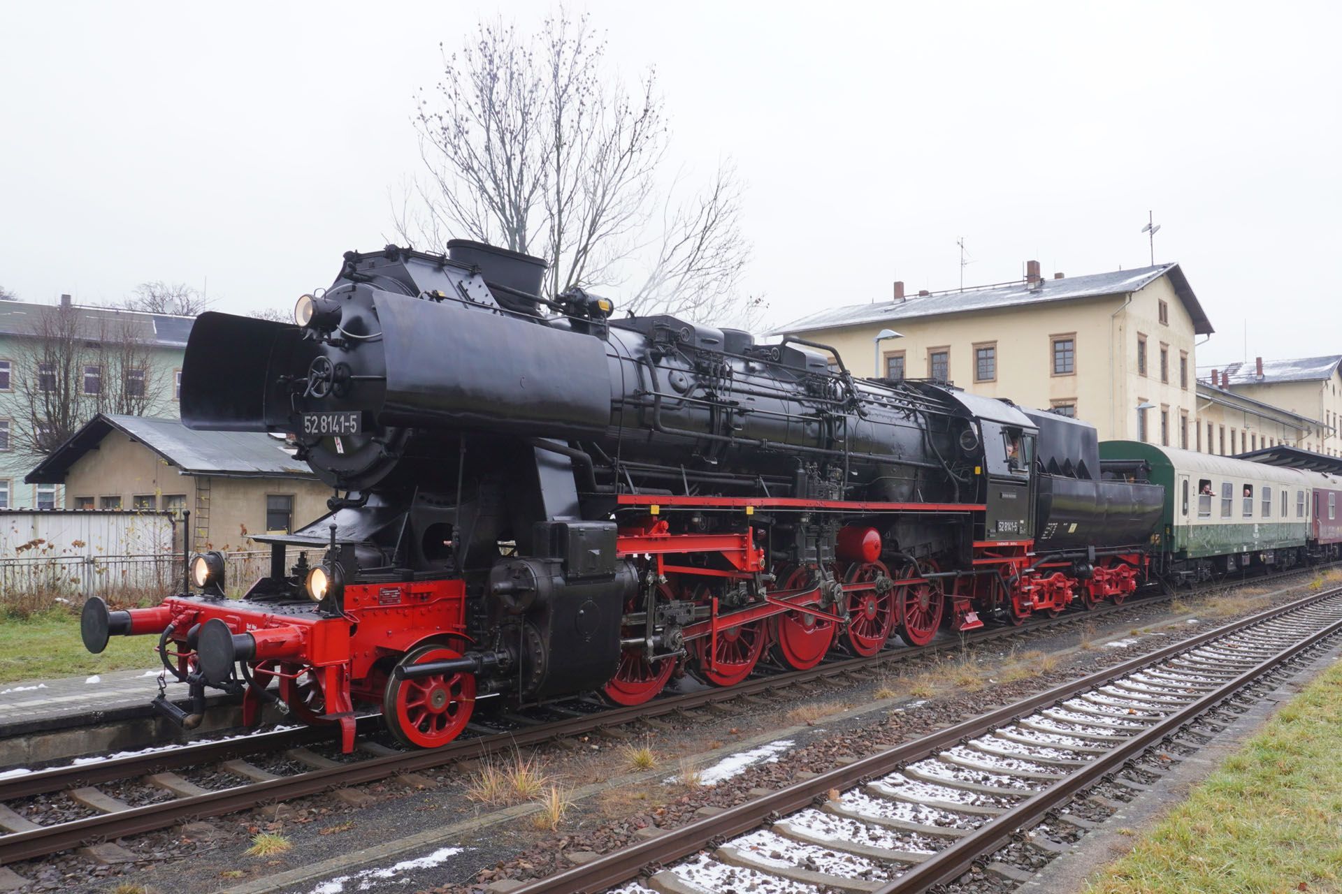 Black steam locomotive with red wheels pulling green passenger cars on wet railway tracks