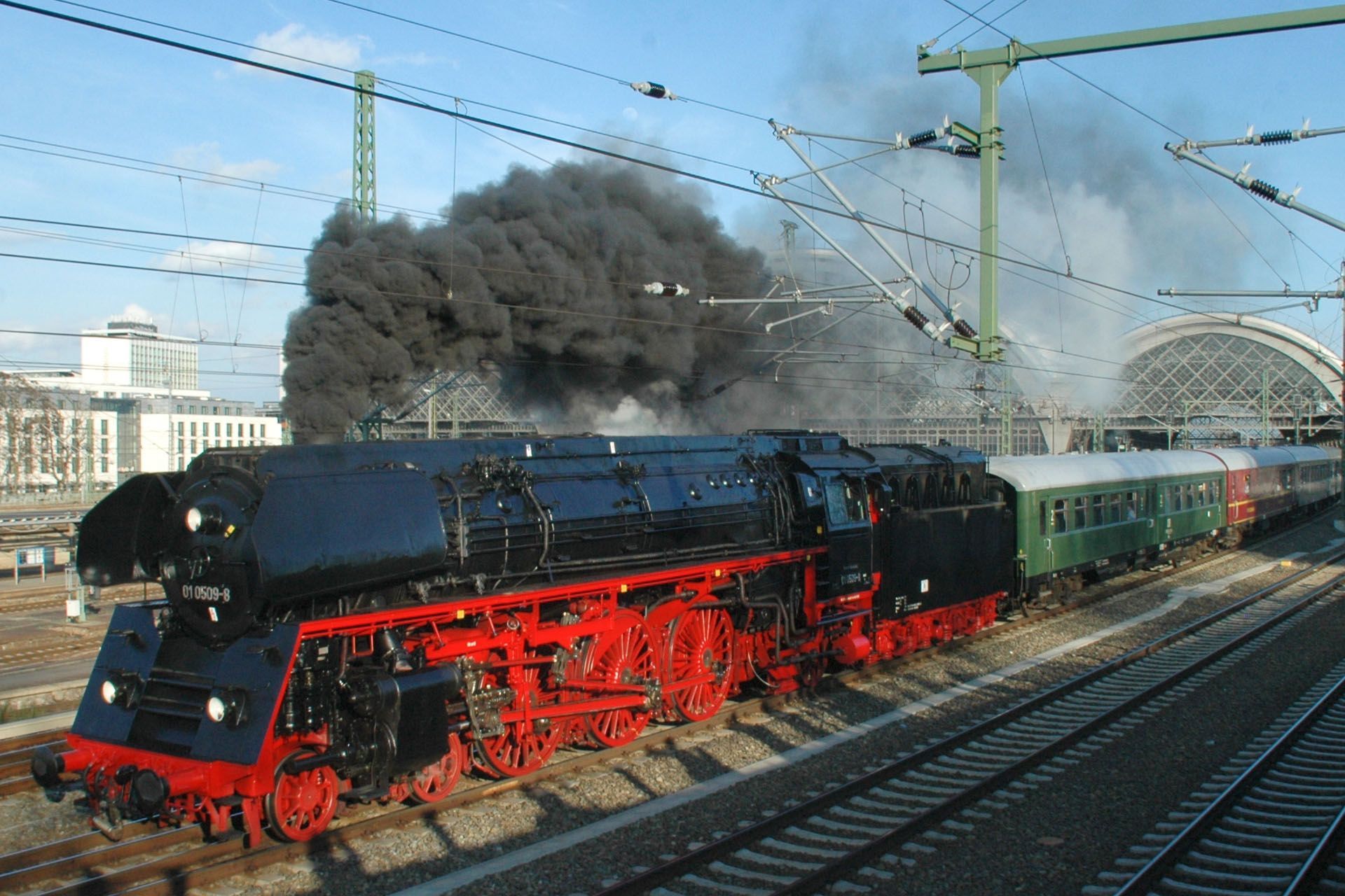 Black steam locomotive with red wheels pulling green passenger cars, billowing smoke on railway tracks