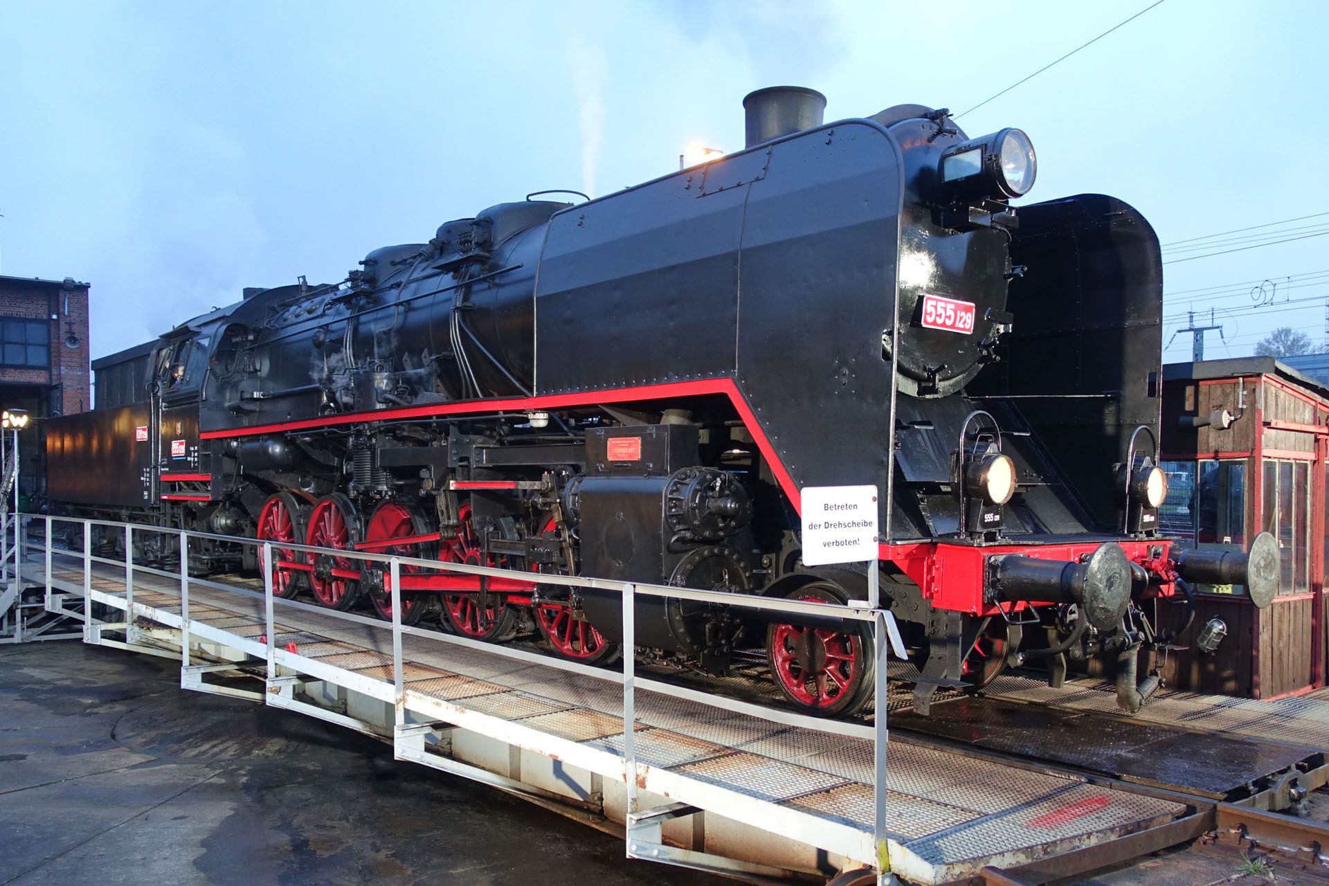 Black steam locomotive on tracks at a rail yard, viewed from the side behind a white railing.