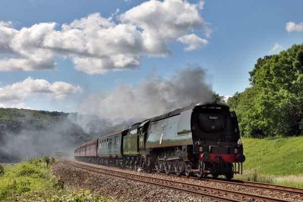 Steam train traveling on tracks through a green, hilly landscape.