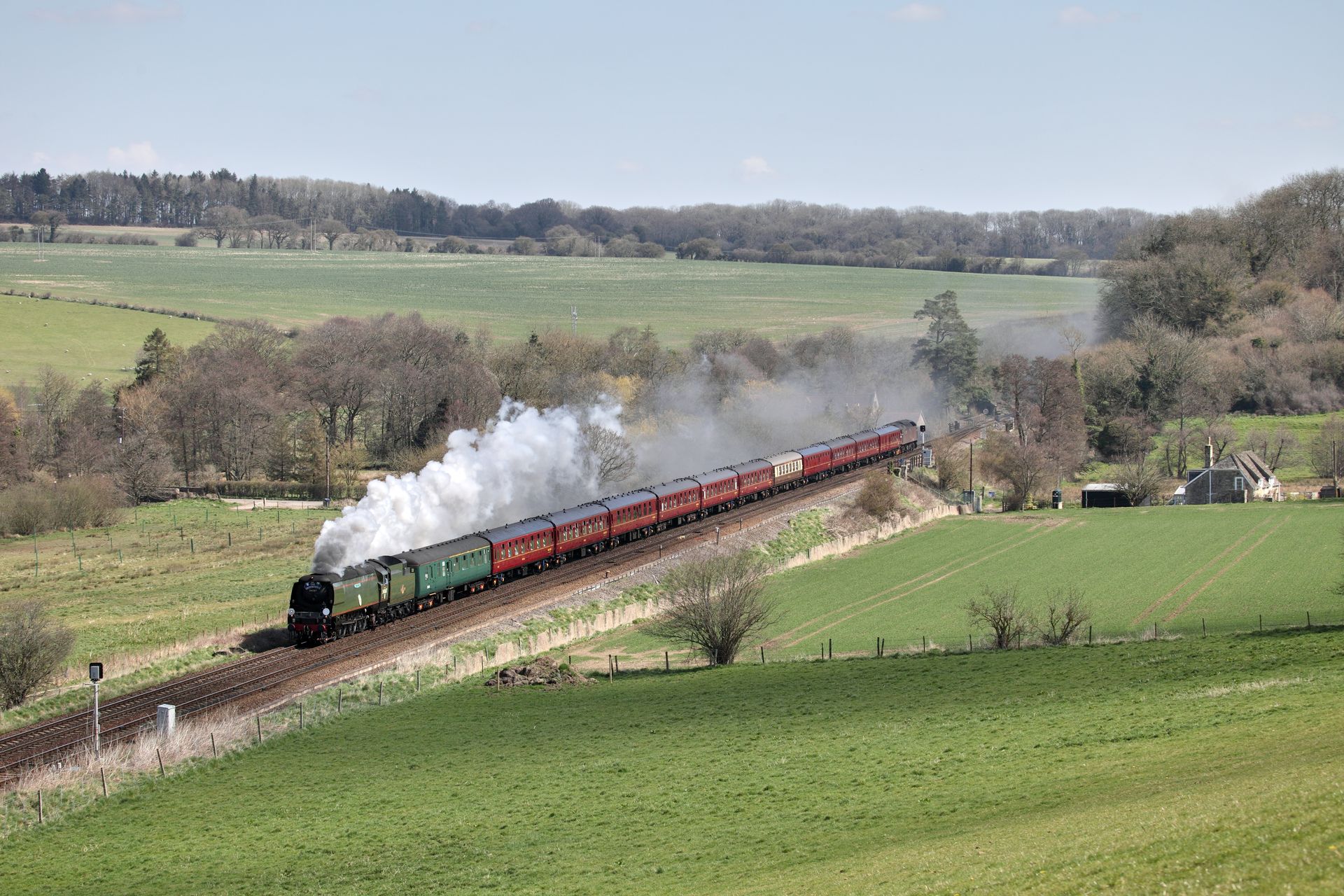 Steam train traveling along tracks through a hilly, grassy landscape, passing under a stone bridge.