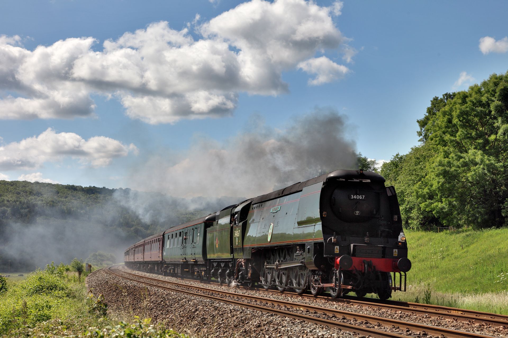 Steam train traveling through a rural landscape, emerging from a tunnel. Green and red carriages.