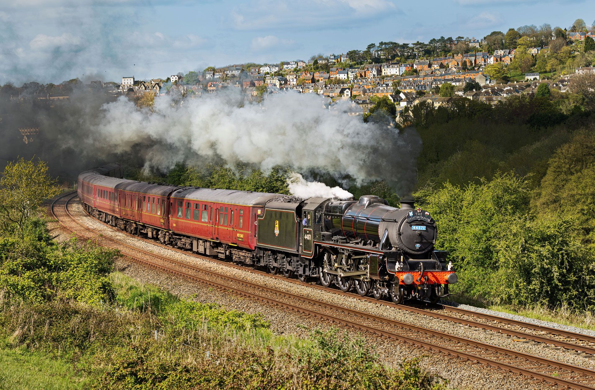 Steam locomotive pulling red passenger cars through a green hillside landscape with smoke billowing behind