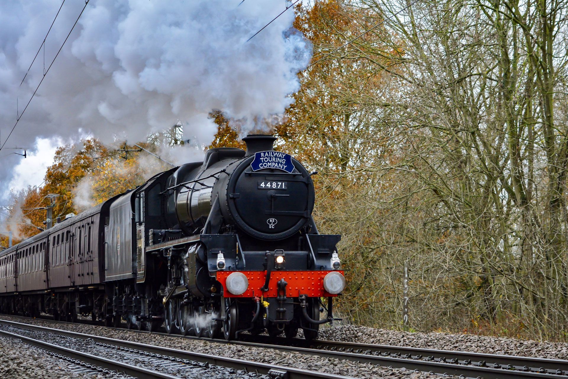Steam locomotive pulling a passenger train through a wooded countryside, with smoke billowing overhead