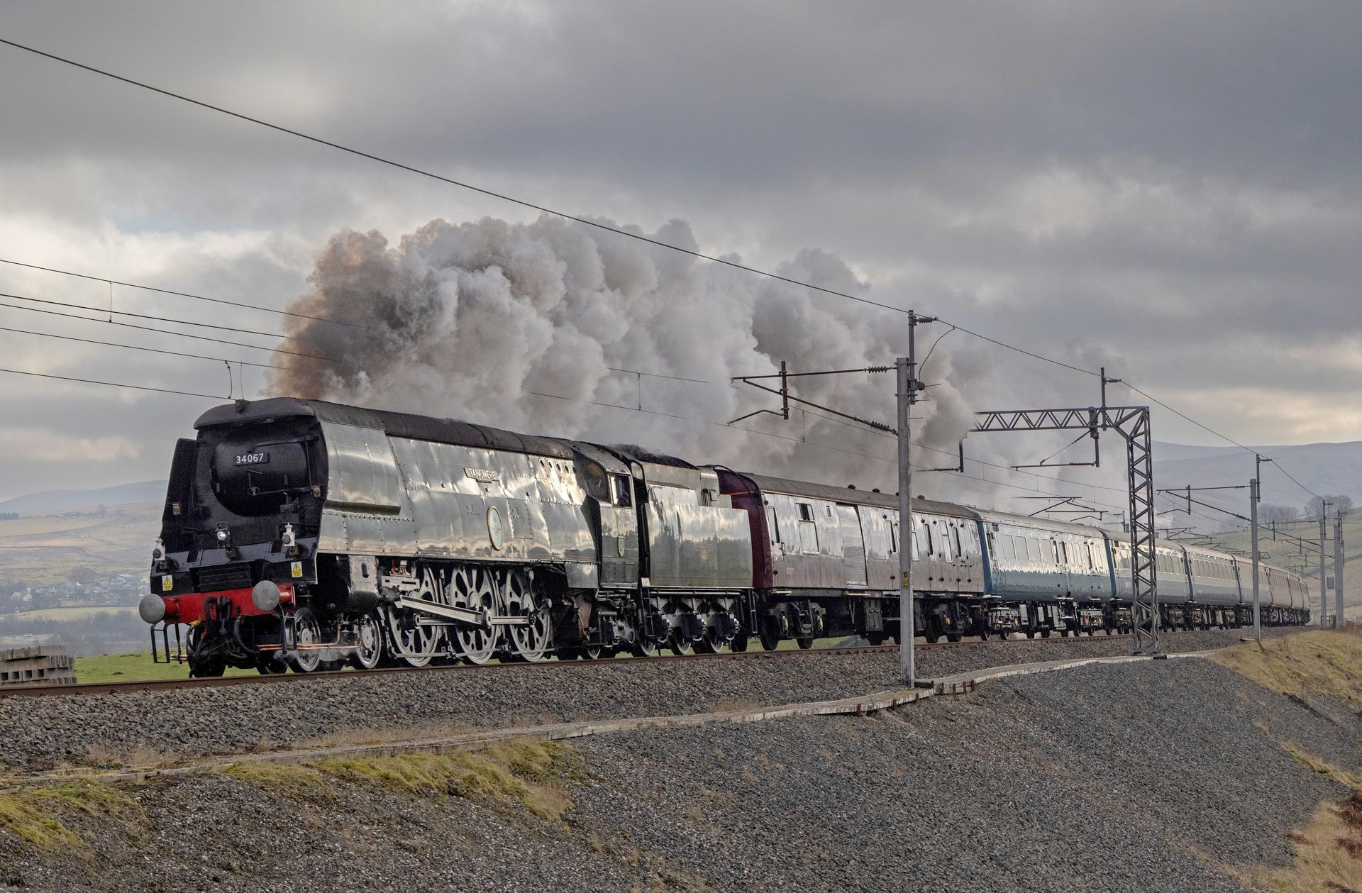 Steam locomotive pulling freight cars along a rural track under a cloudy sky