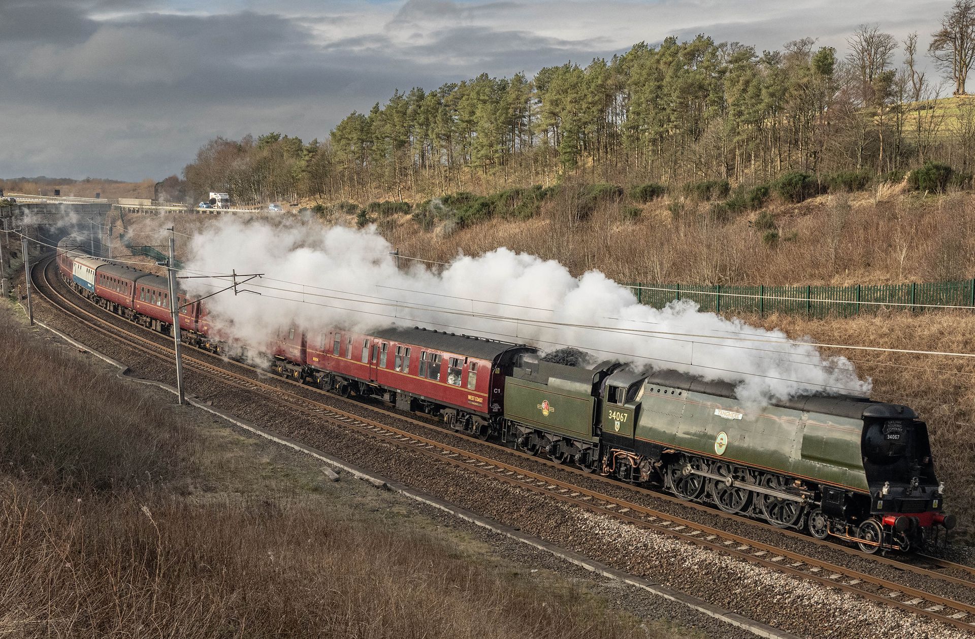 Steam train hauling red carriages through a countryside curve, billowing white smoke under a cloudy sky