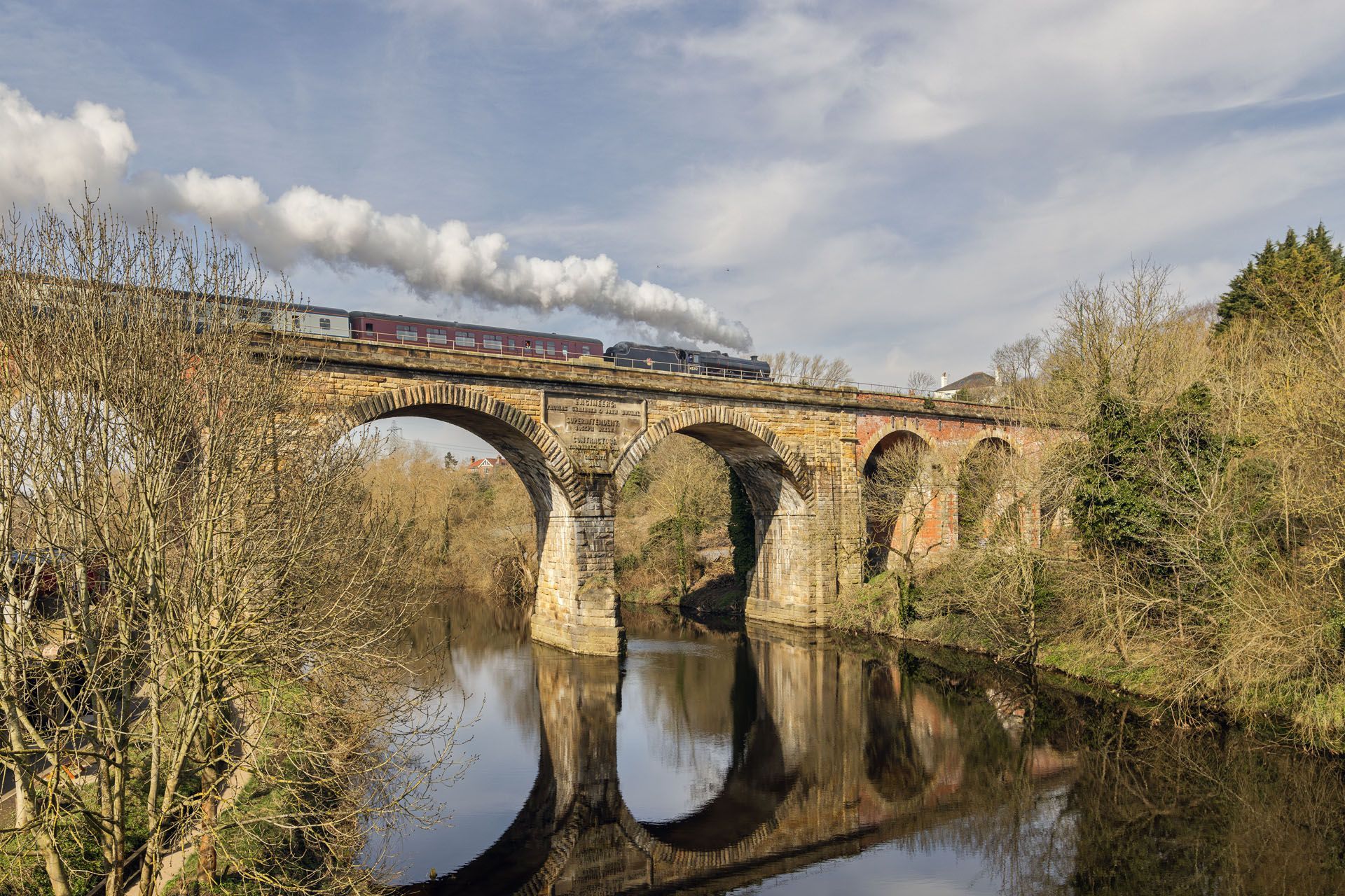Stone arched bridge over a calm river, with autumn trees and reflections under a cloudy sky