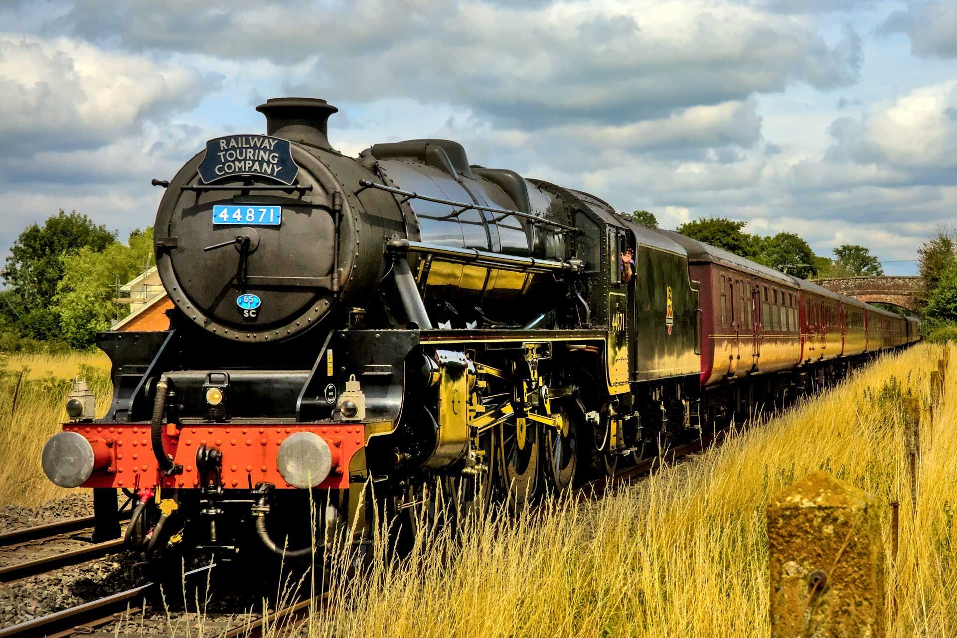 A black steam locomotive travels along tracks through a grassy field under a cloudy blue sky.
