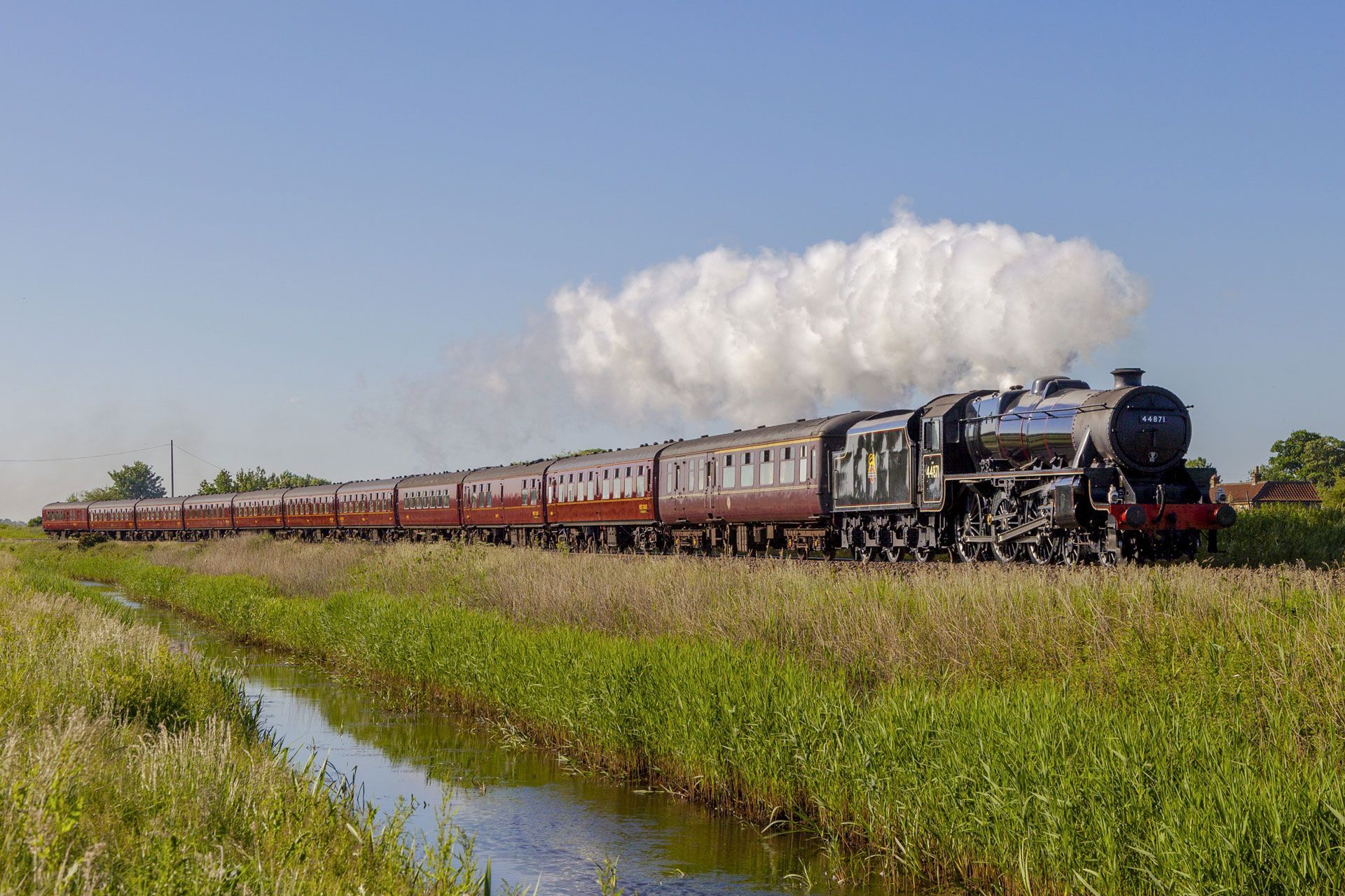 Steam locomotive pulling a long passenger train through green fields under a blue sky