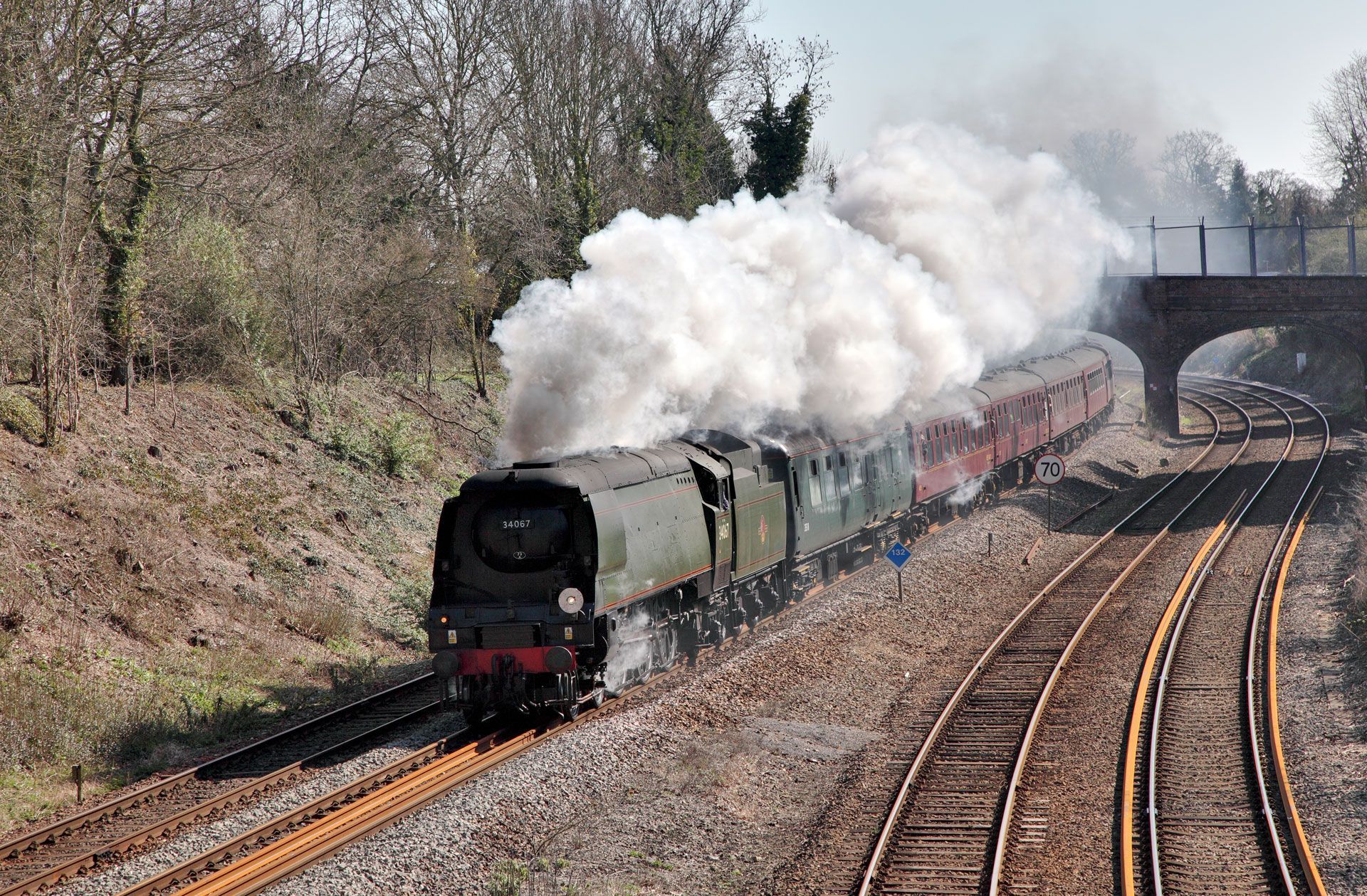 Steam train curving through a wooded railway cutting, trailing thick white smoke under a bridge