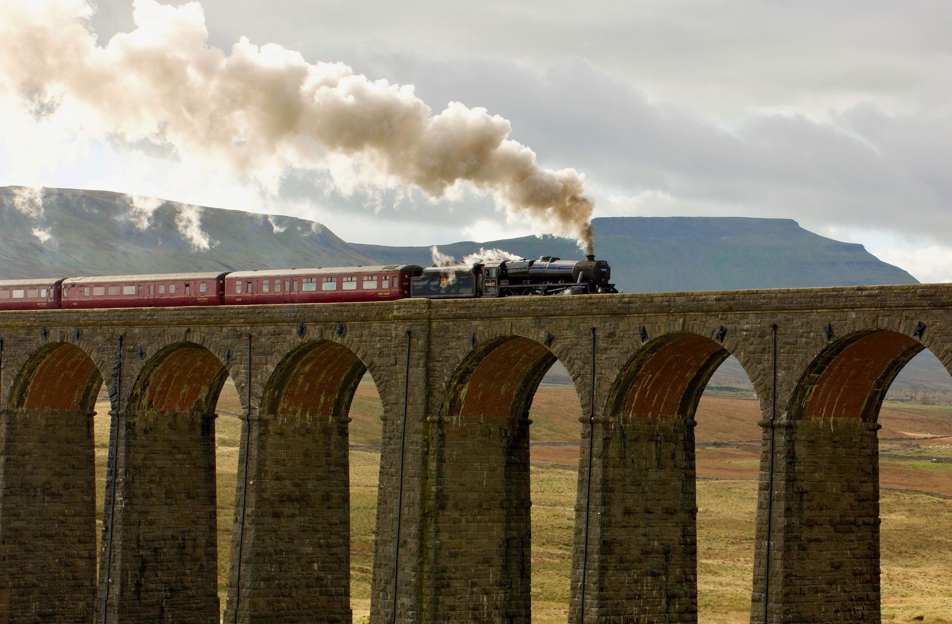 Steam train crossing a stone viaduct, pulling red carriages under a cloudy sky