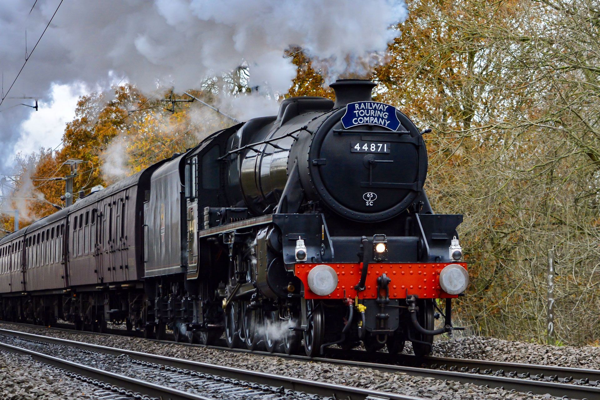 Black steam locomotive pulling passenger cars through a wooded curve, with billowing smoke.