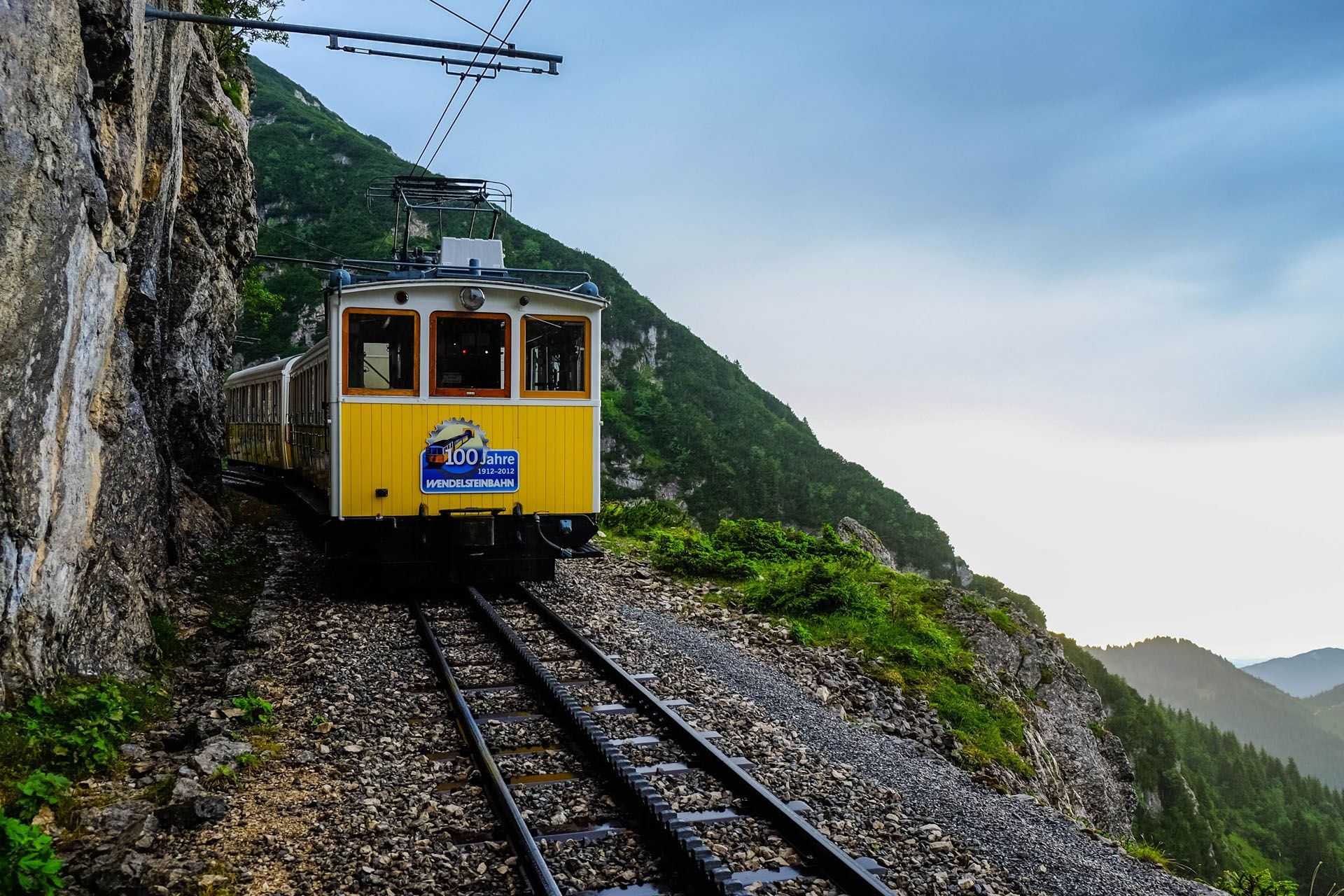 Yellow cable car ascending a mountain rail track along a cliffside; green vegetation, cloudy sky.