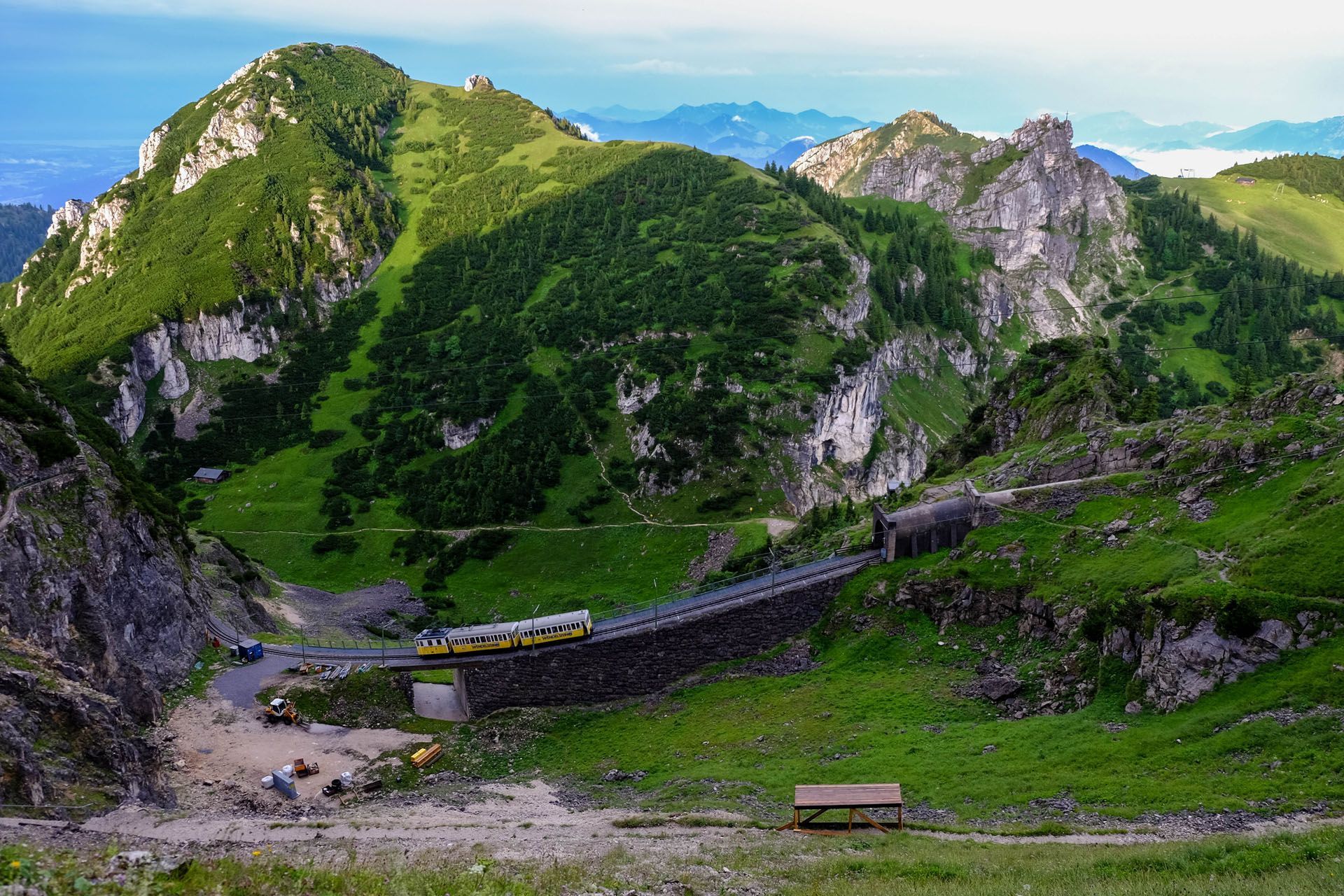 Mountain landscape with a train traversing a stone bridge, green hills, and rocky peaks.