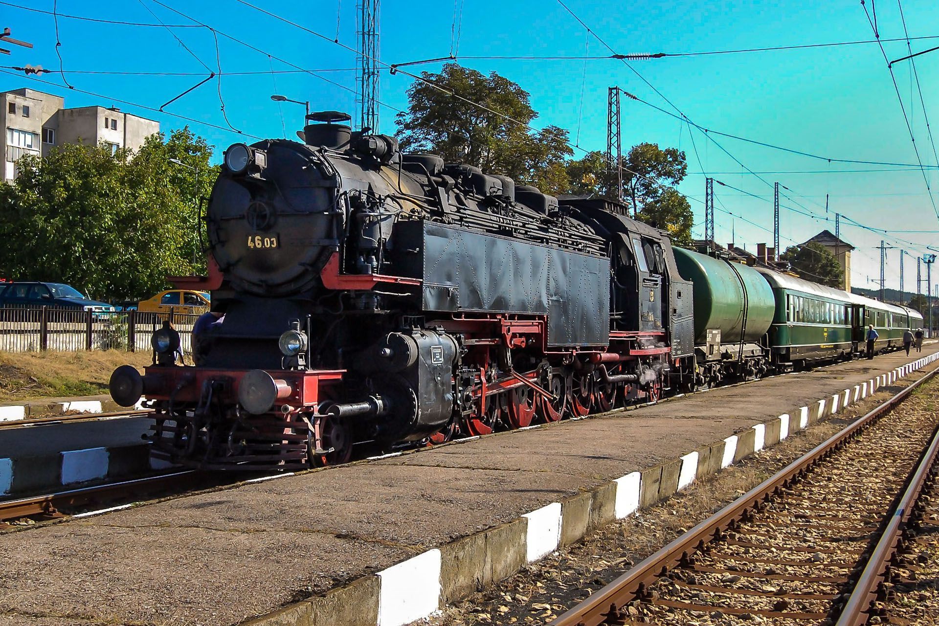 Black steam locomotive pulling green cars on railway tracks under a bright blue sky.