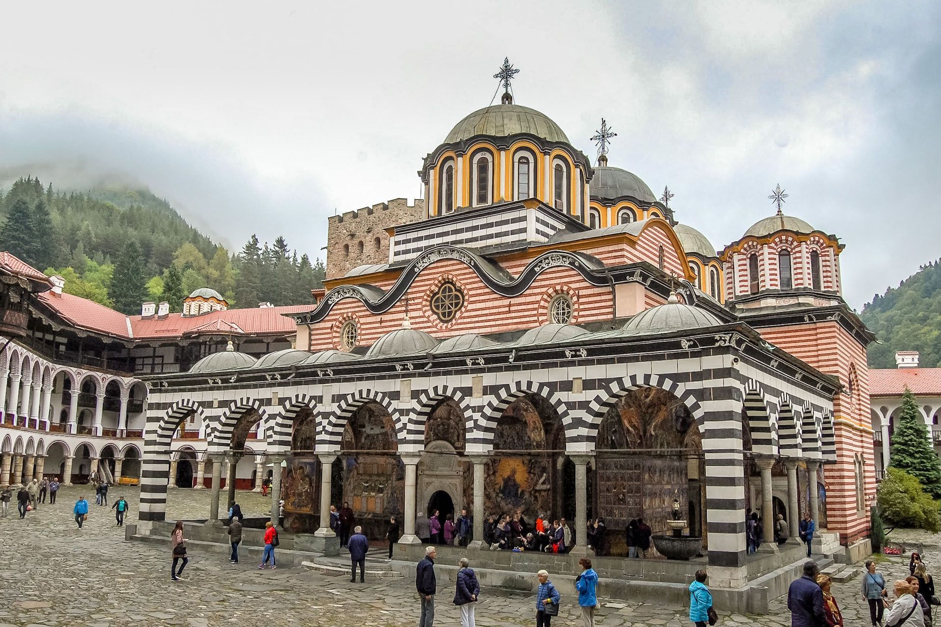 Ornate striped stone church with domes in a mountain square, people gathered outside.