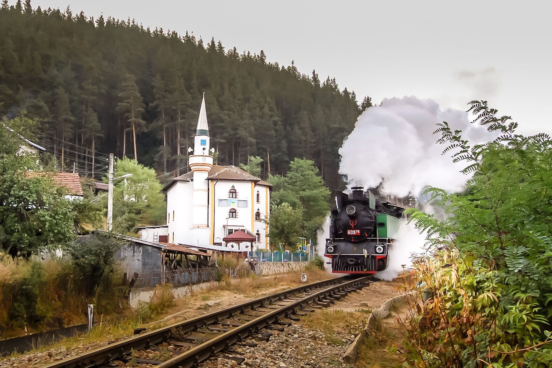 Steam locomotive on tracks beside a small church, with white smoke in a forested mountain valley.