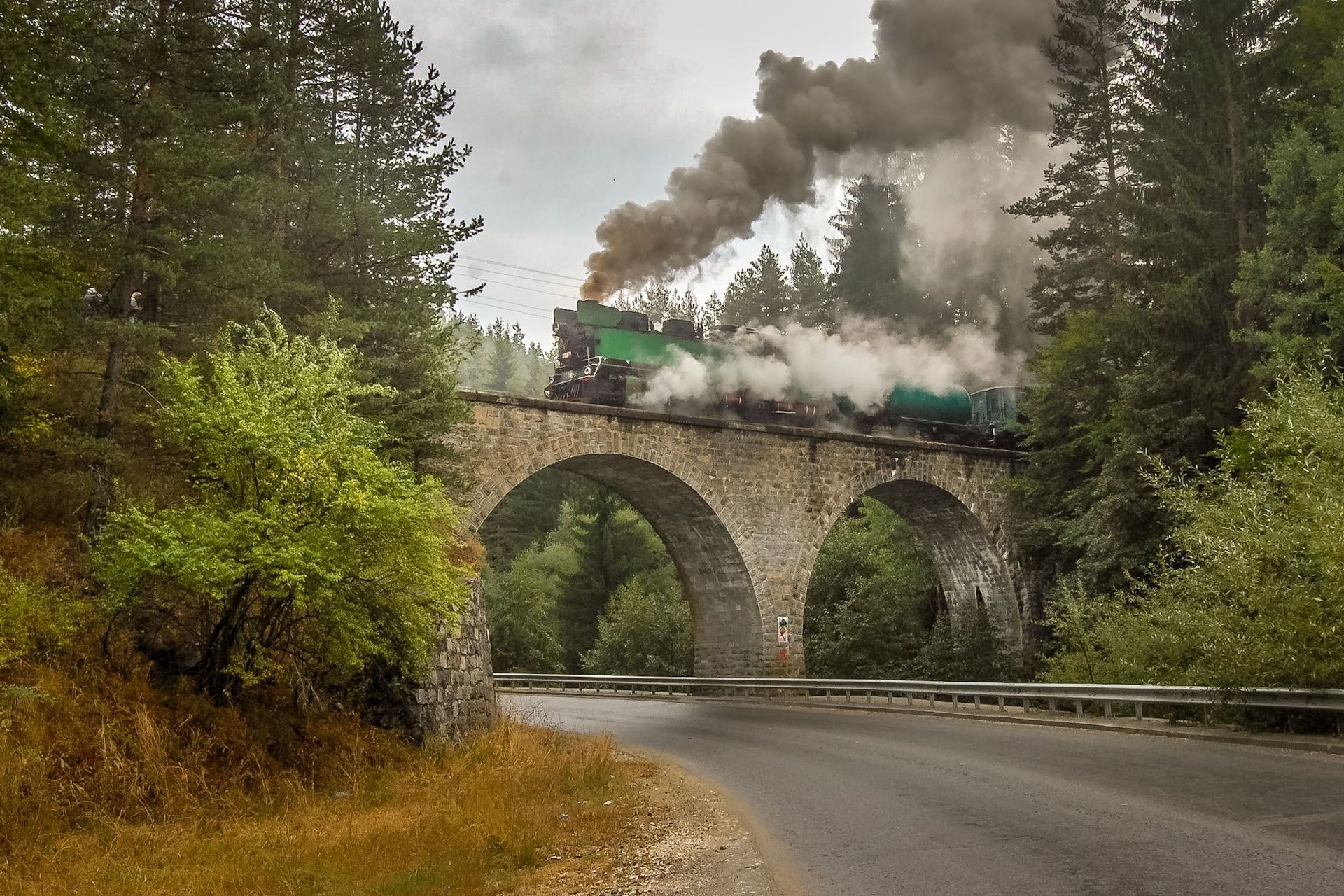 Steam locomotive crossing stone viaduct over a forest road, with smoke billowing into the gray sky