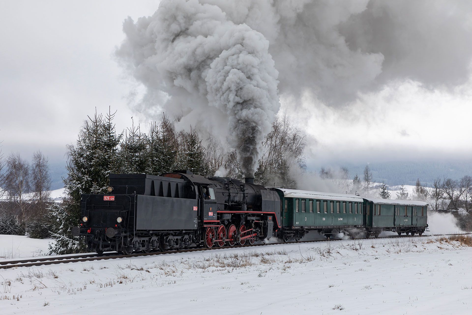 Steam locomotive pulling green cars through snowy landscape, billowing white smoke