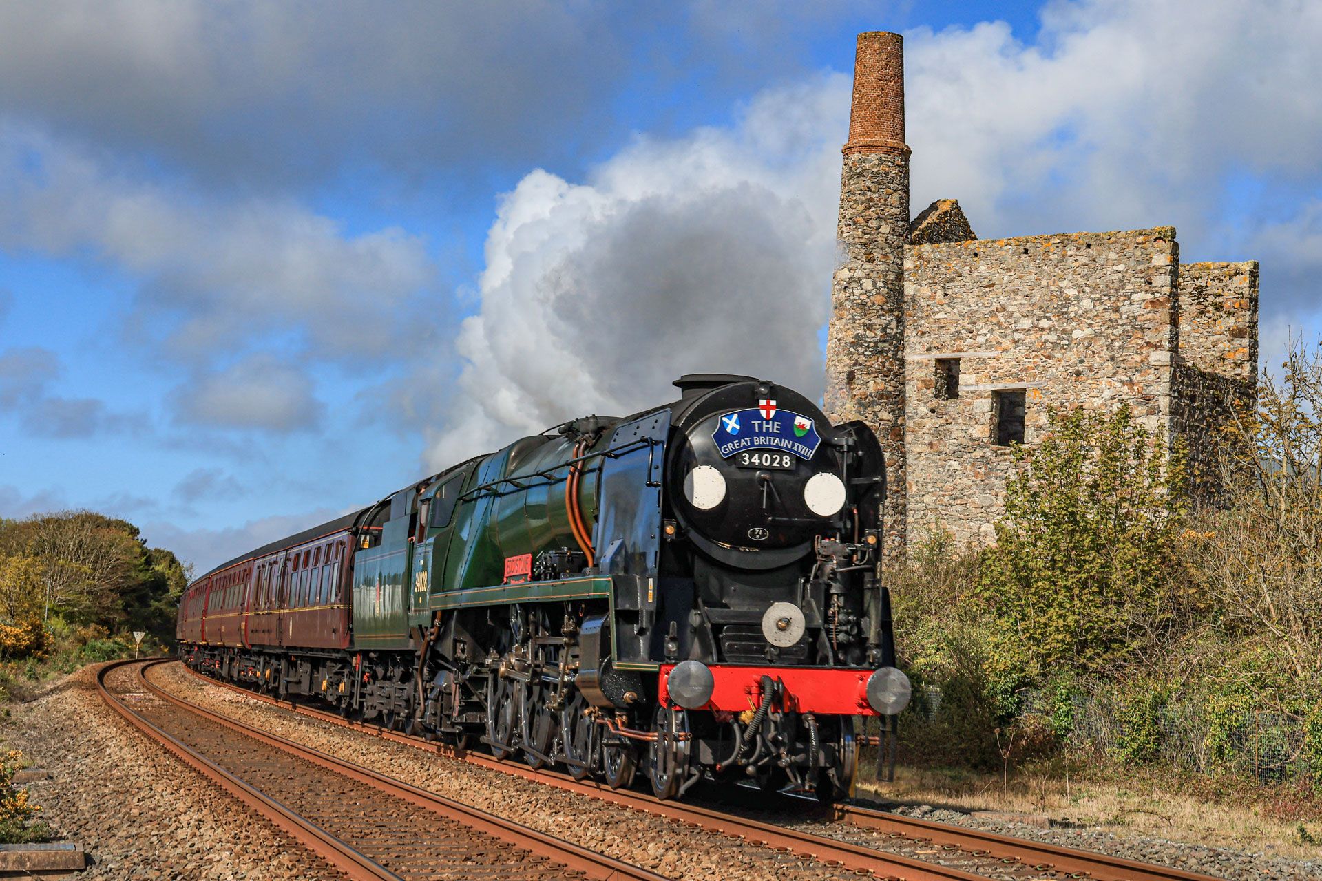 Steam locomotive pulling green passenger cars past a stone building under a cloudy sky