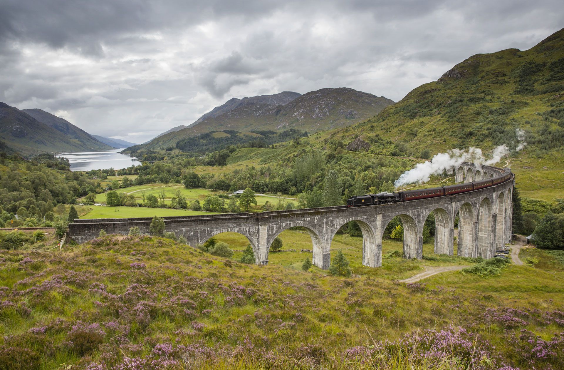 Steam train crossing a stone viaduct in a lush green mountain valley under cloudy skies
