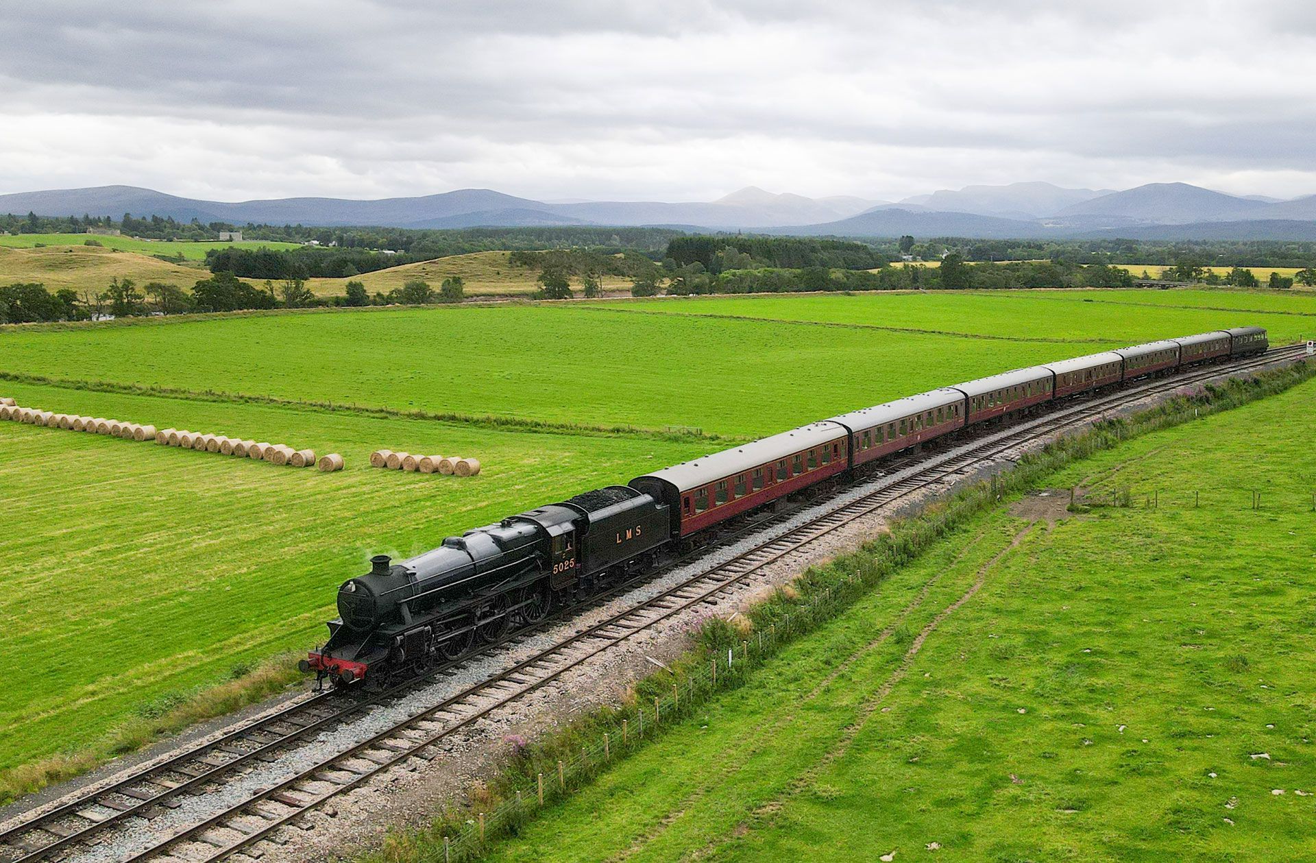 Black steam train with red cars crossing a green countryside under cloudy skies