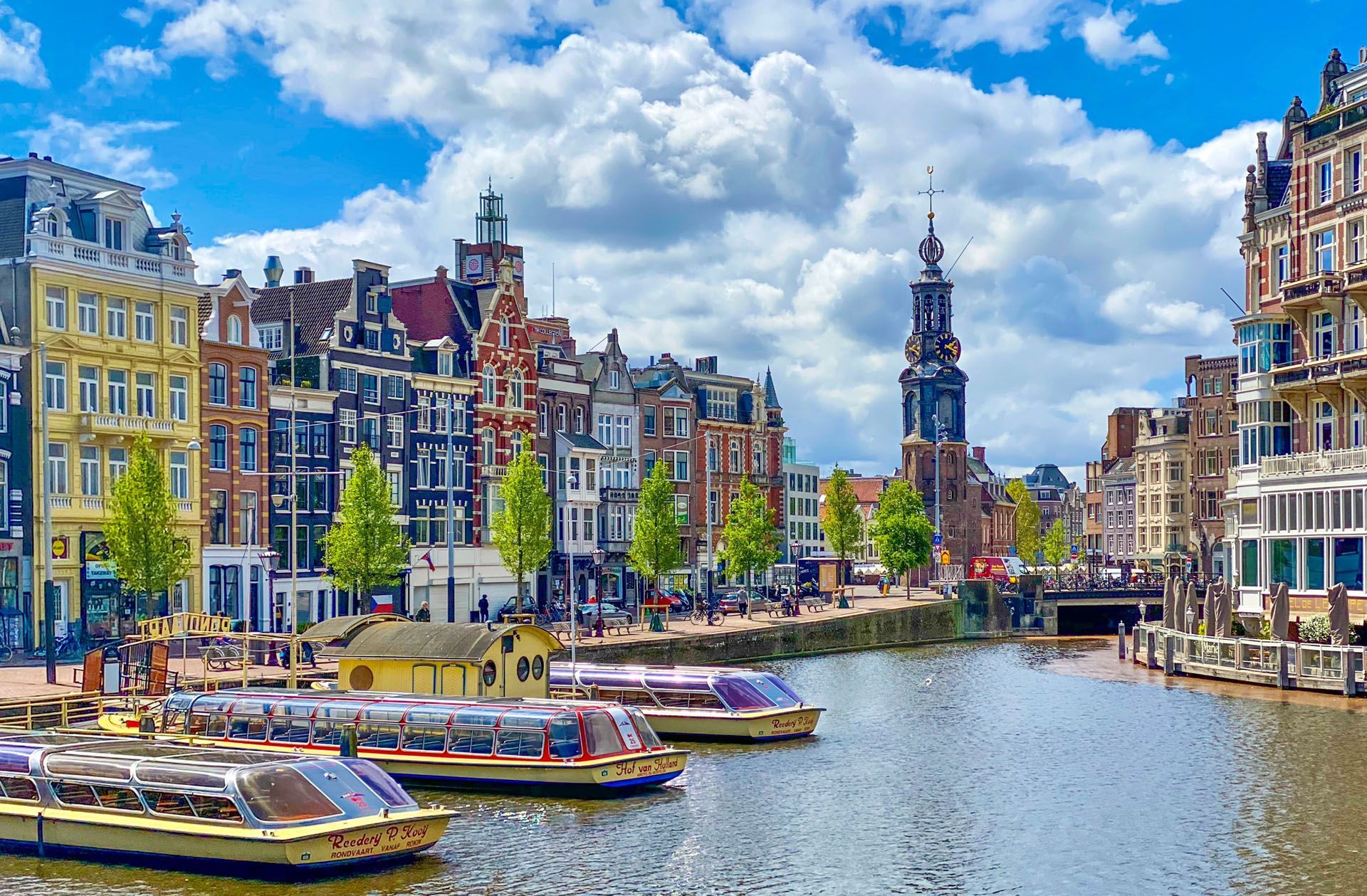 Canal in Amsterdam with colorful narrow buildings, boats, and a church tower under a bright blue sky