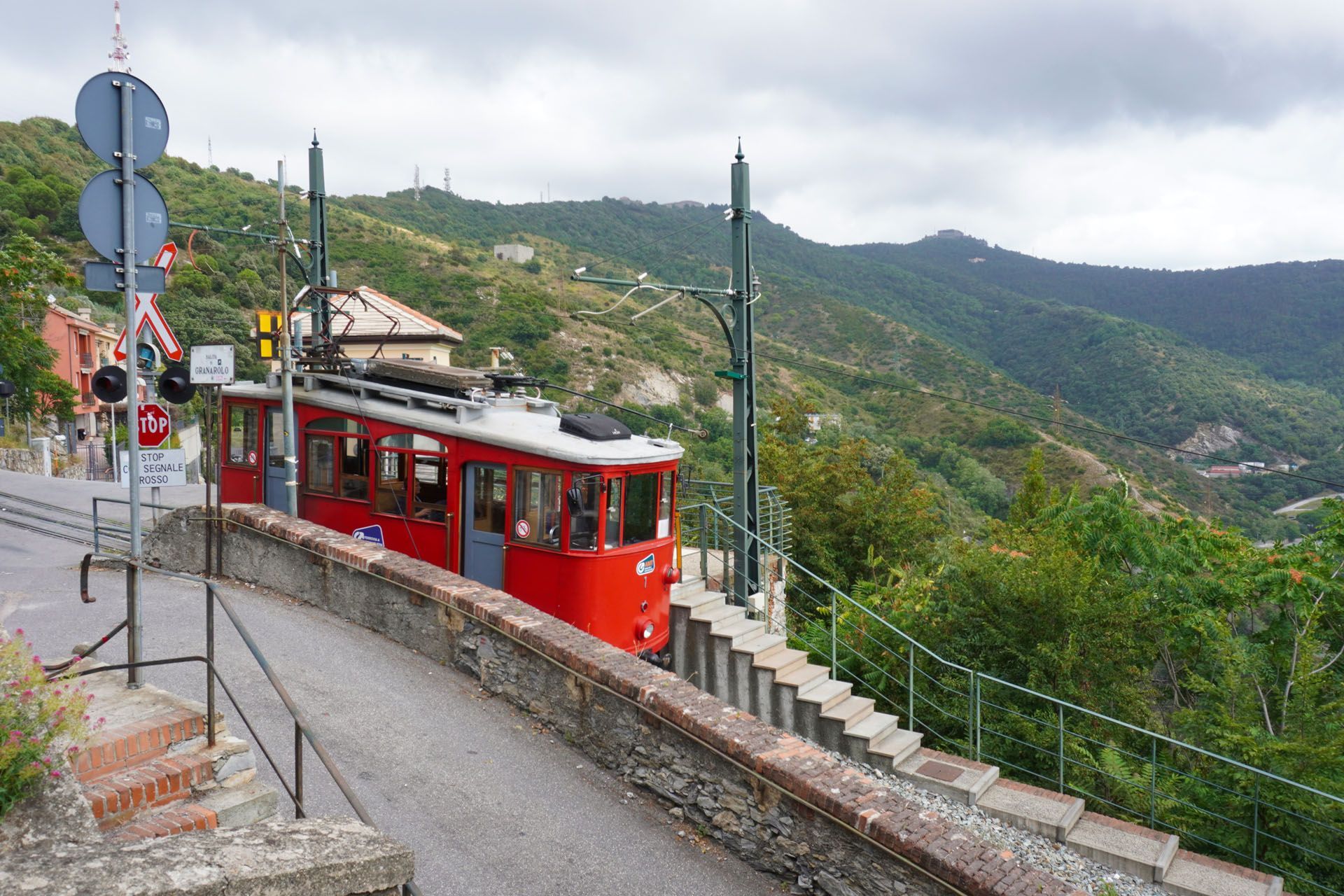 Red funicular car on tracks, traveling up a hillside with green trees and mountains in the background.