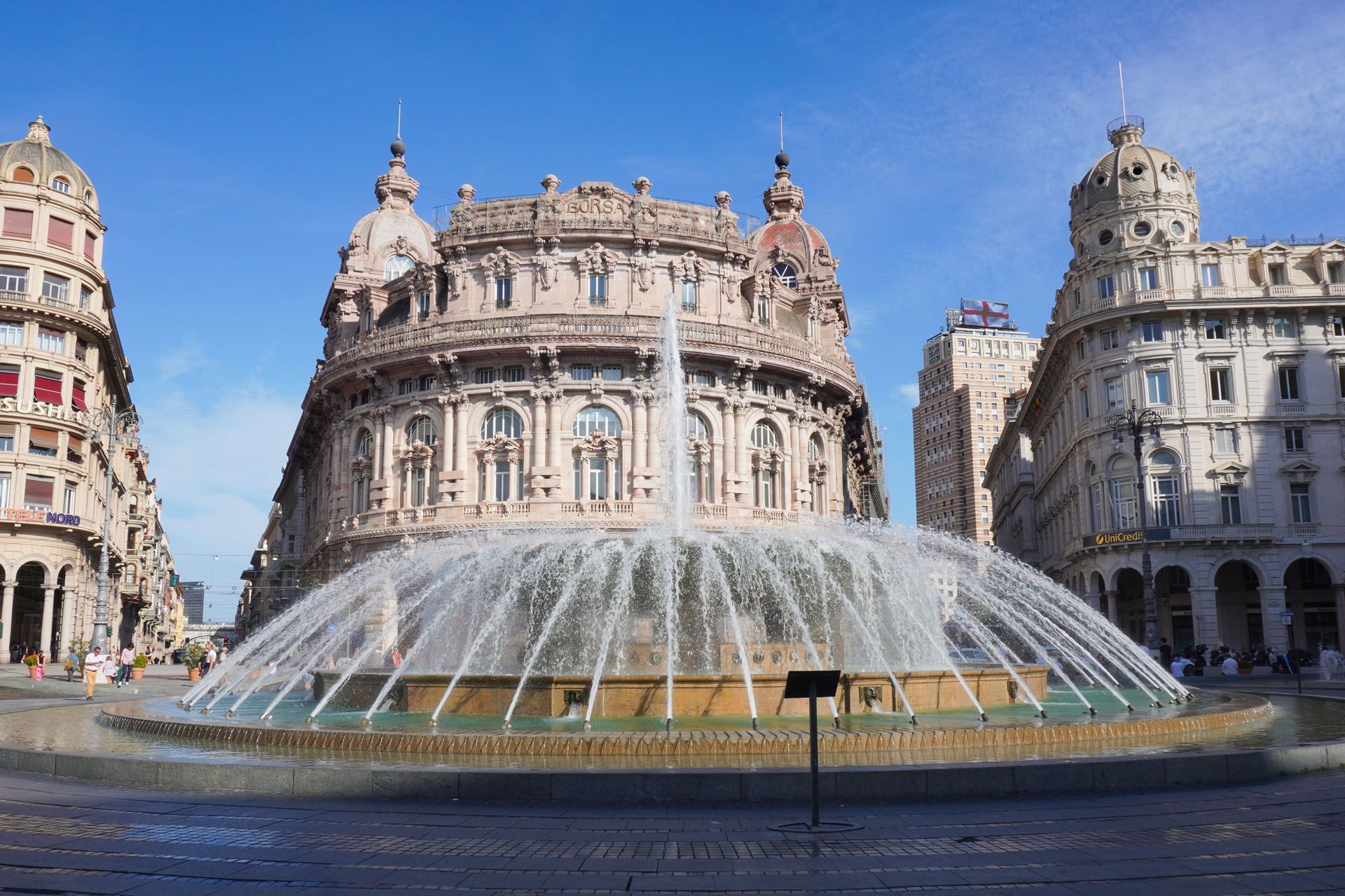 Fountain in a plaza with a large, ornate building in Genoa, Italy. Blue sky, water spraying.