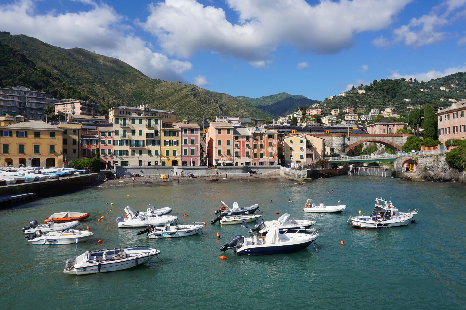 Boats in turquoise water, colorful buildings along a harbor, green hills in background.