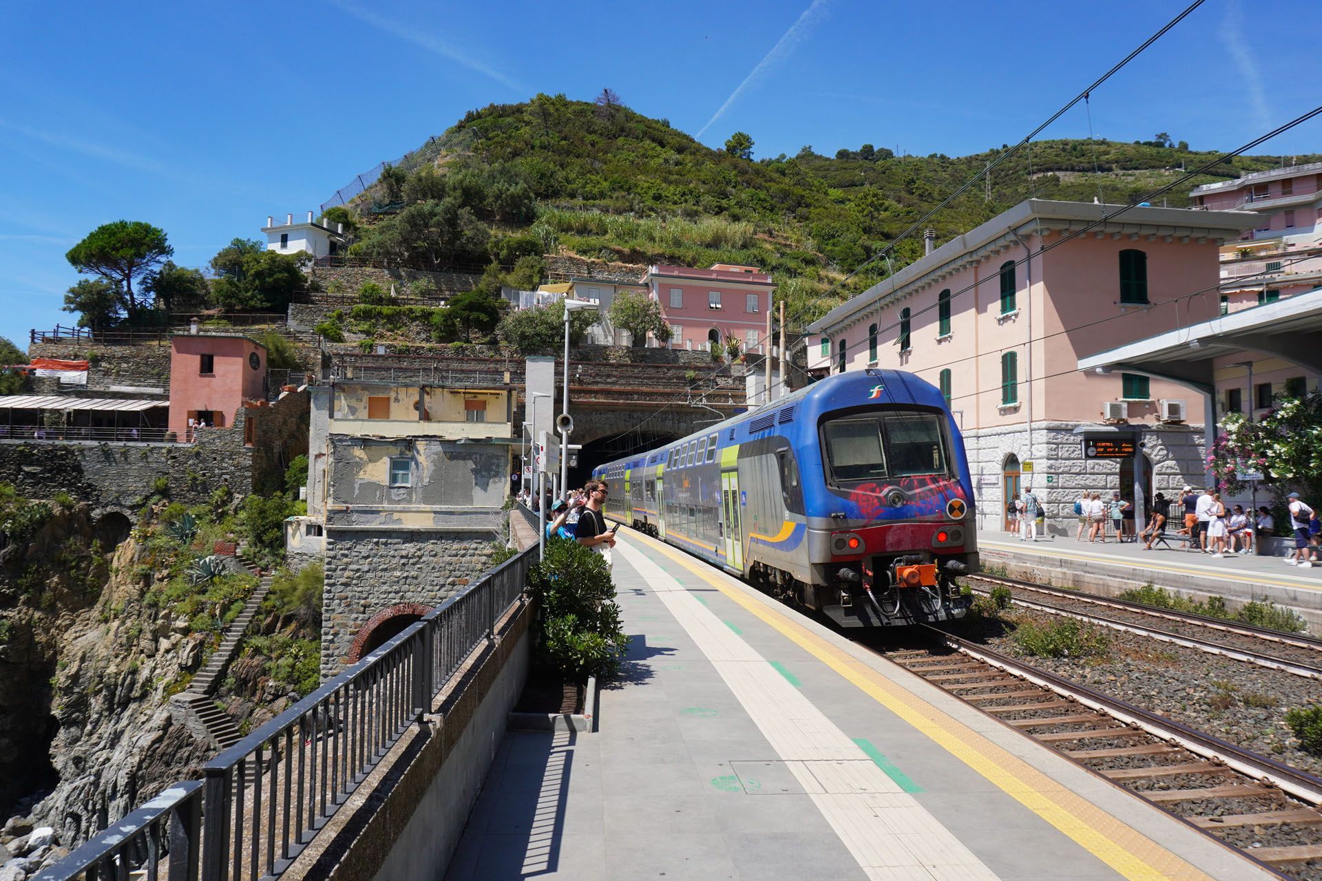 Train pulling into station with hillside town in the background under a blue sky.