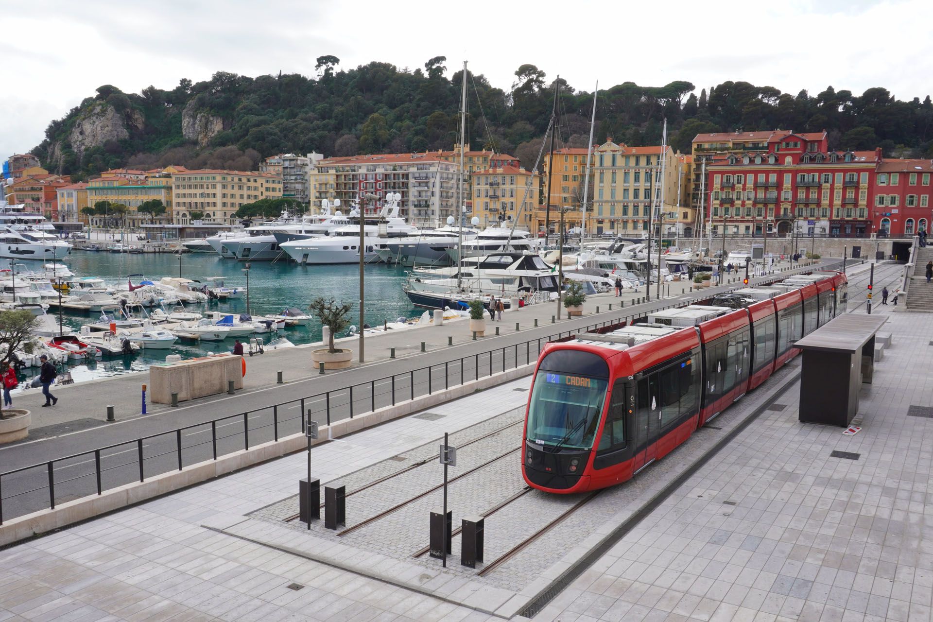 Red tram on tracks by a harbor with boats and buildings.