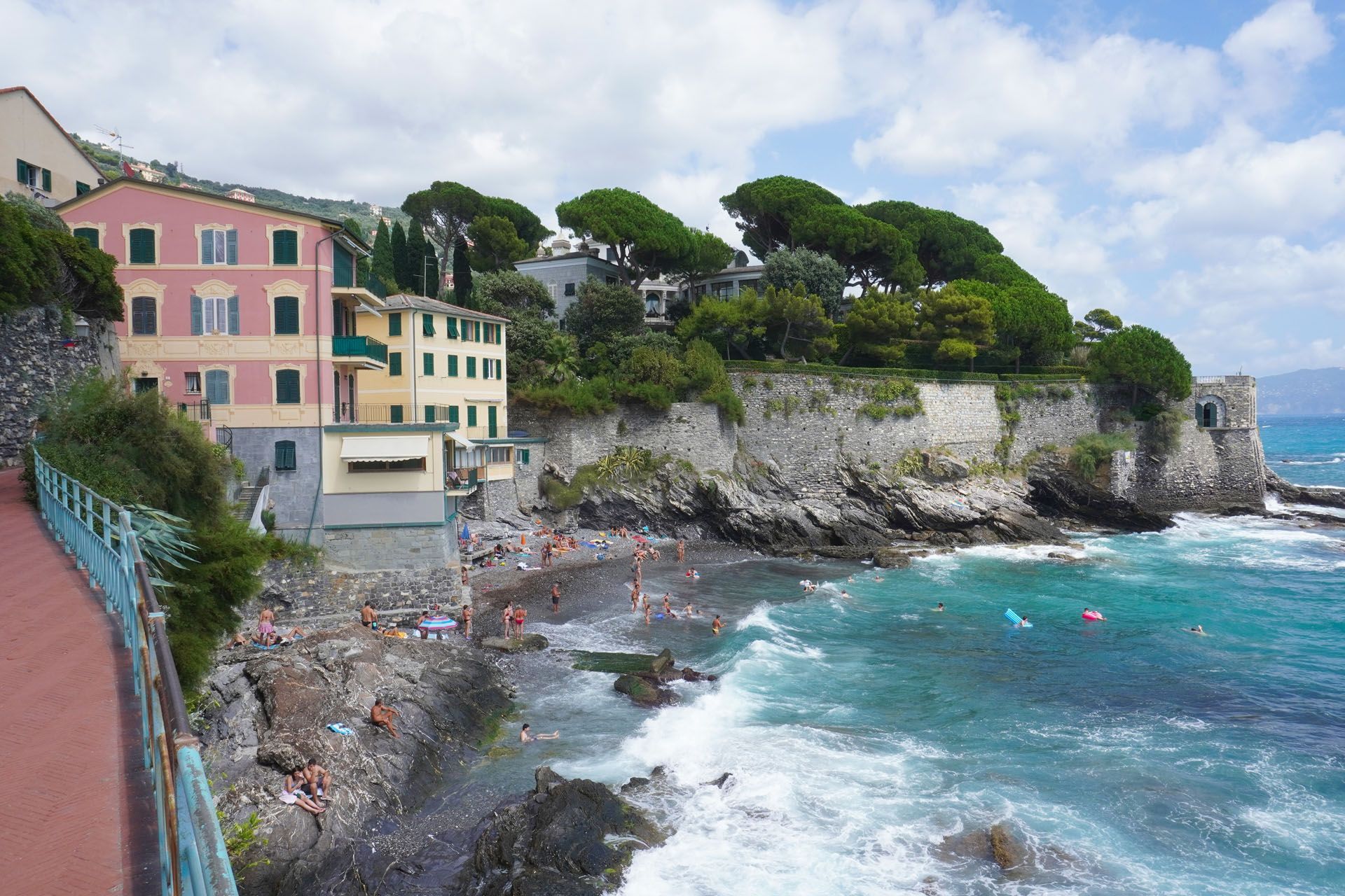 Coastal view: Colorful buildings, rocky shore, turquoise water, people swimming, sunny day.
