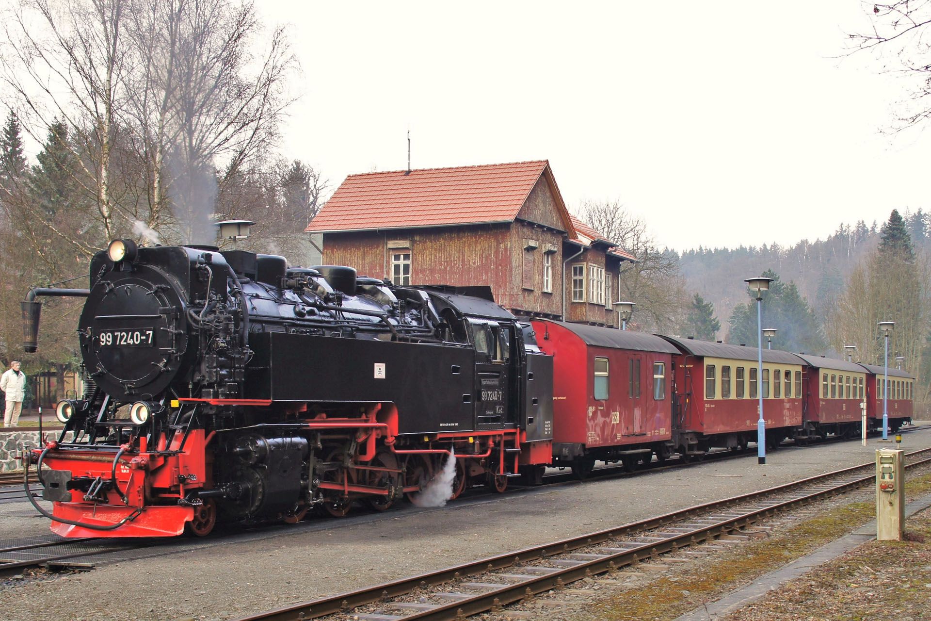 Black steam locomotive pulling red passenger cars past a station on a rainy track