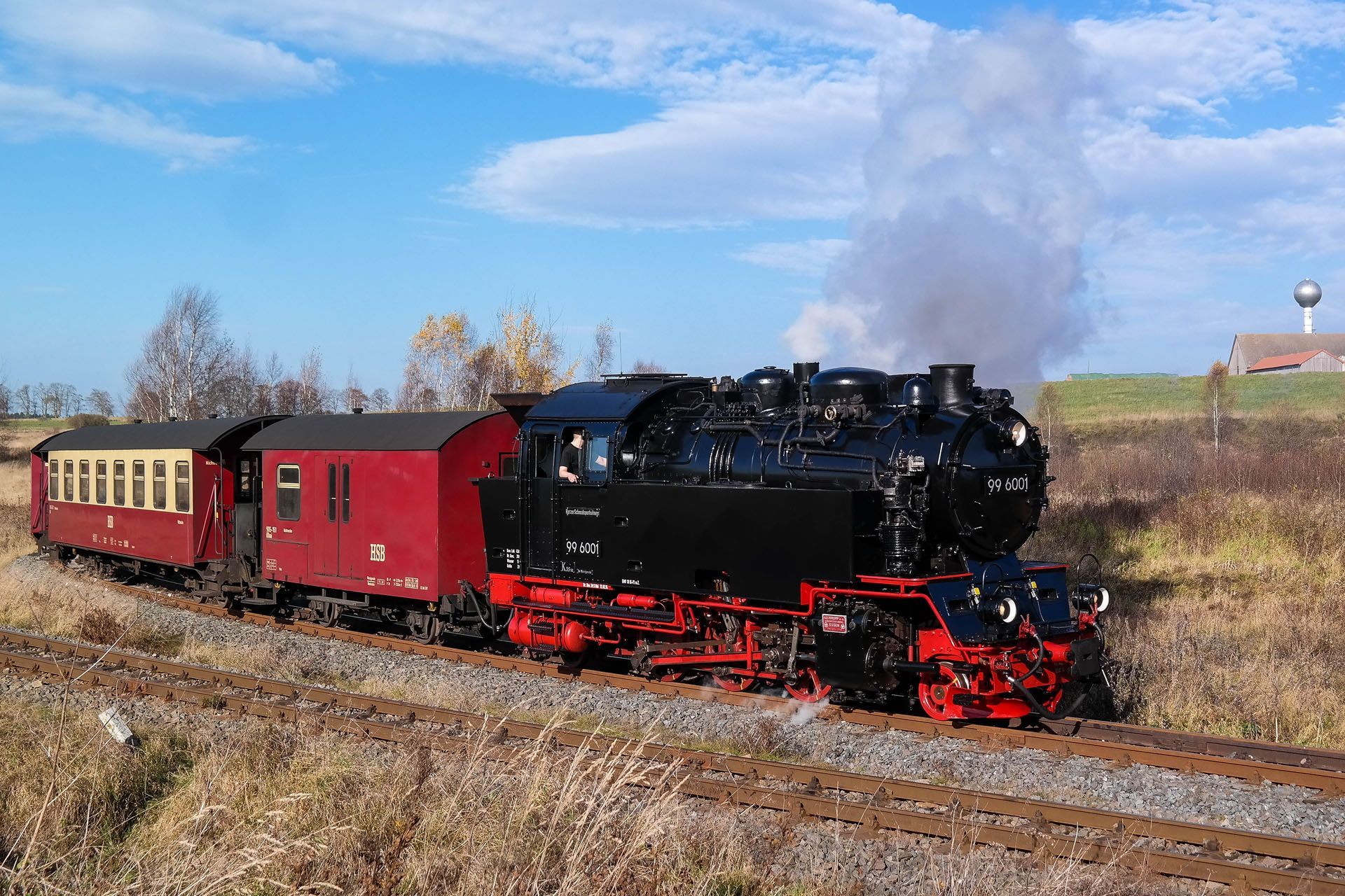 Black steam locomotive pulling red passenger cars through a rural field under a cloudy blue sky