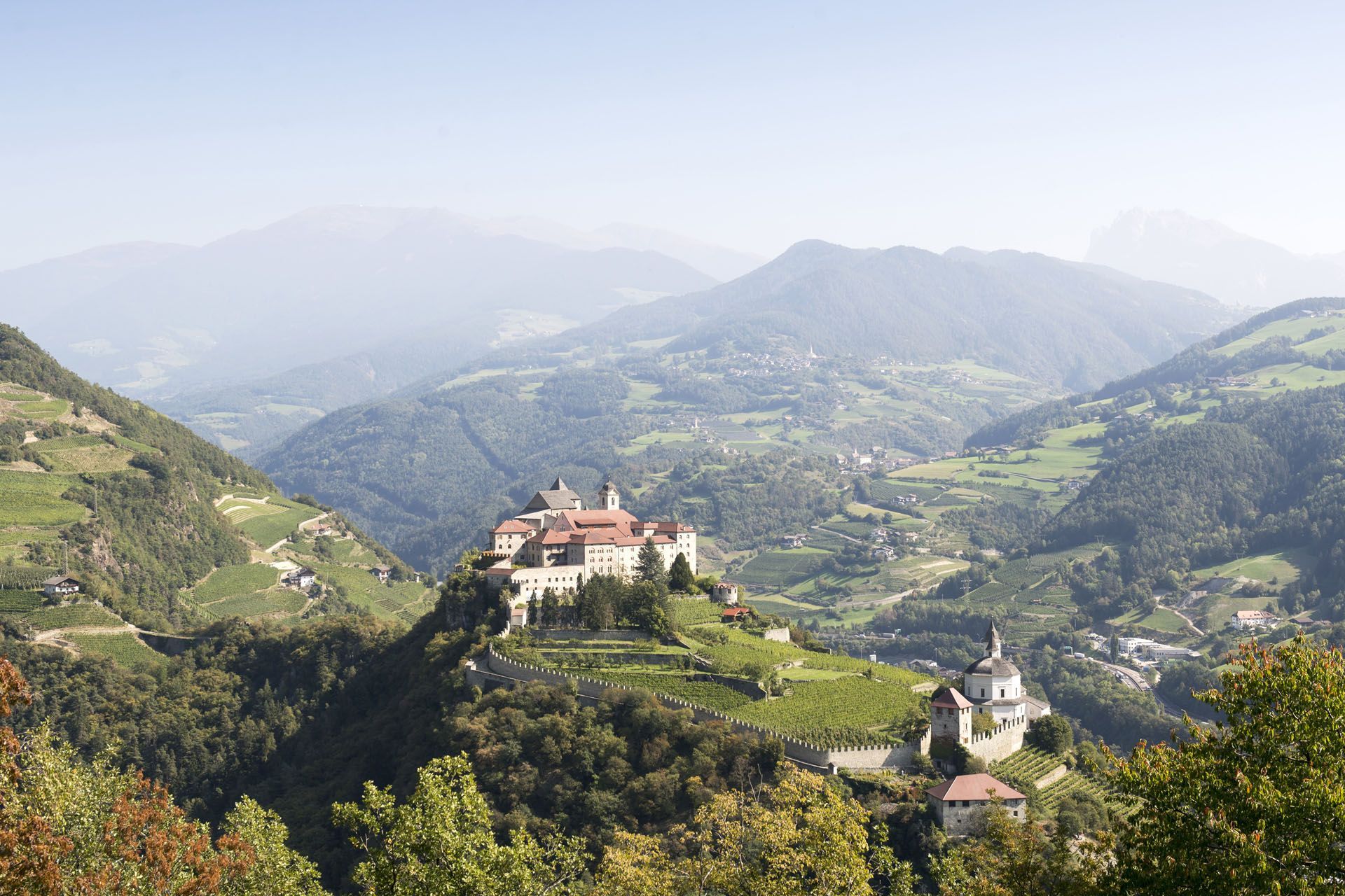 Mountain village with a church and monastery nestled among green hills and hazy mountains.