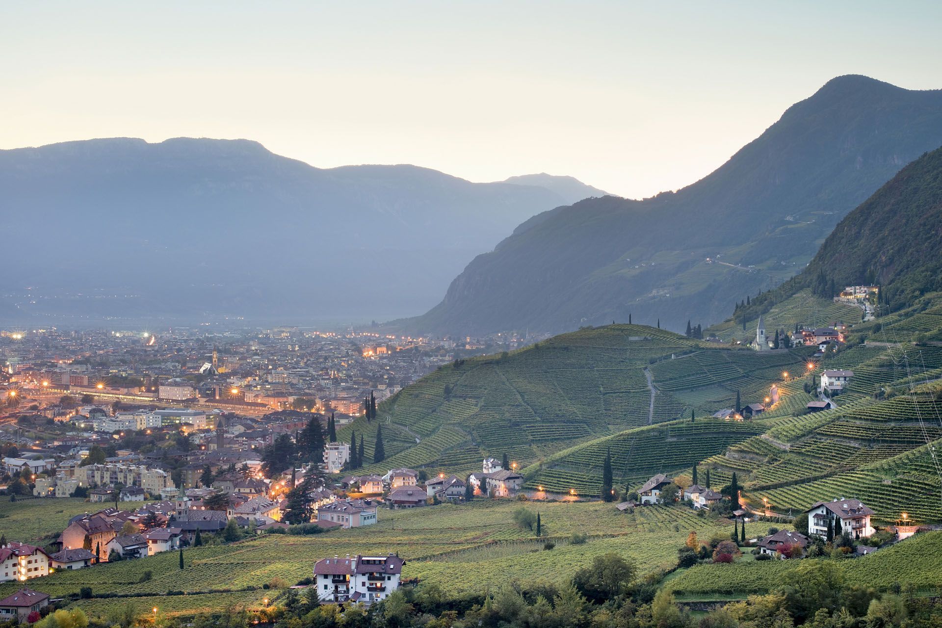 Mountain valley town at dusk with glowing city lights and terraced green hills in the foreground