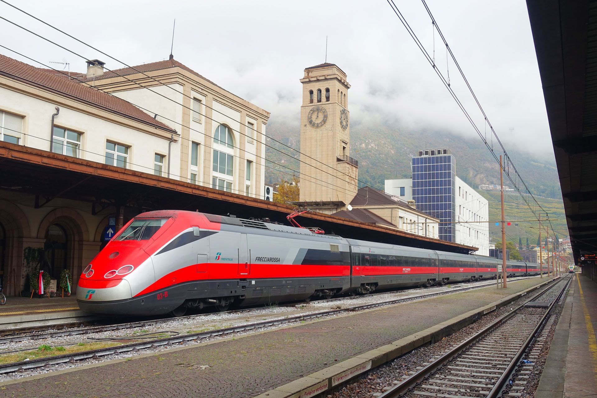 Red and gray high-speed train at a station with mountains and a clock tower in the background
