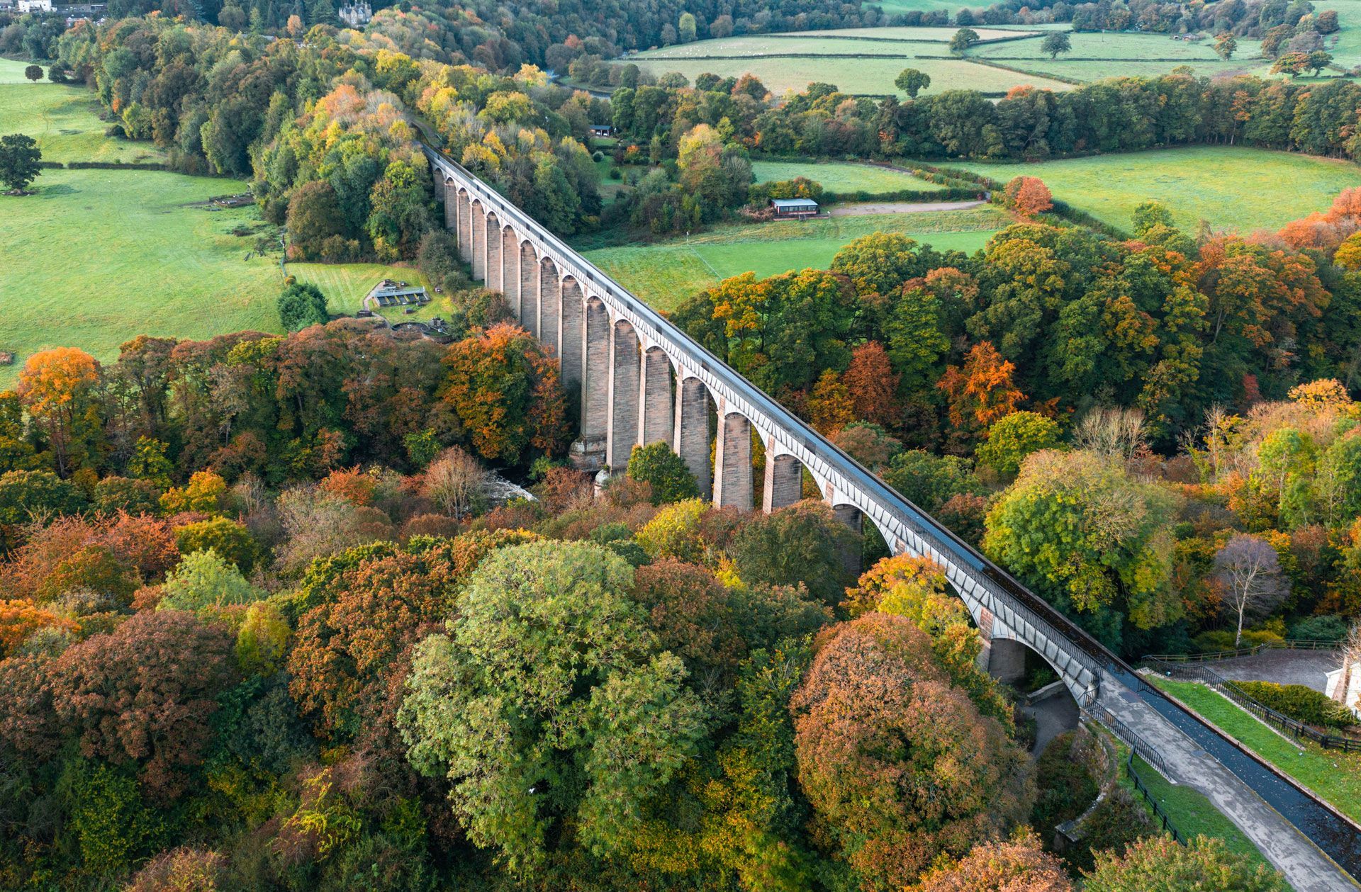 A long stone viaduct crosses a colorful autumn forest and green fields from an aerial view