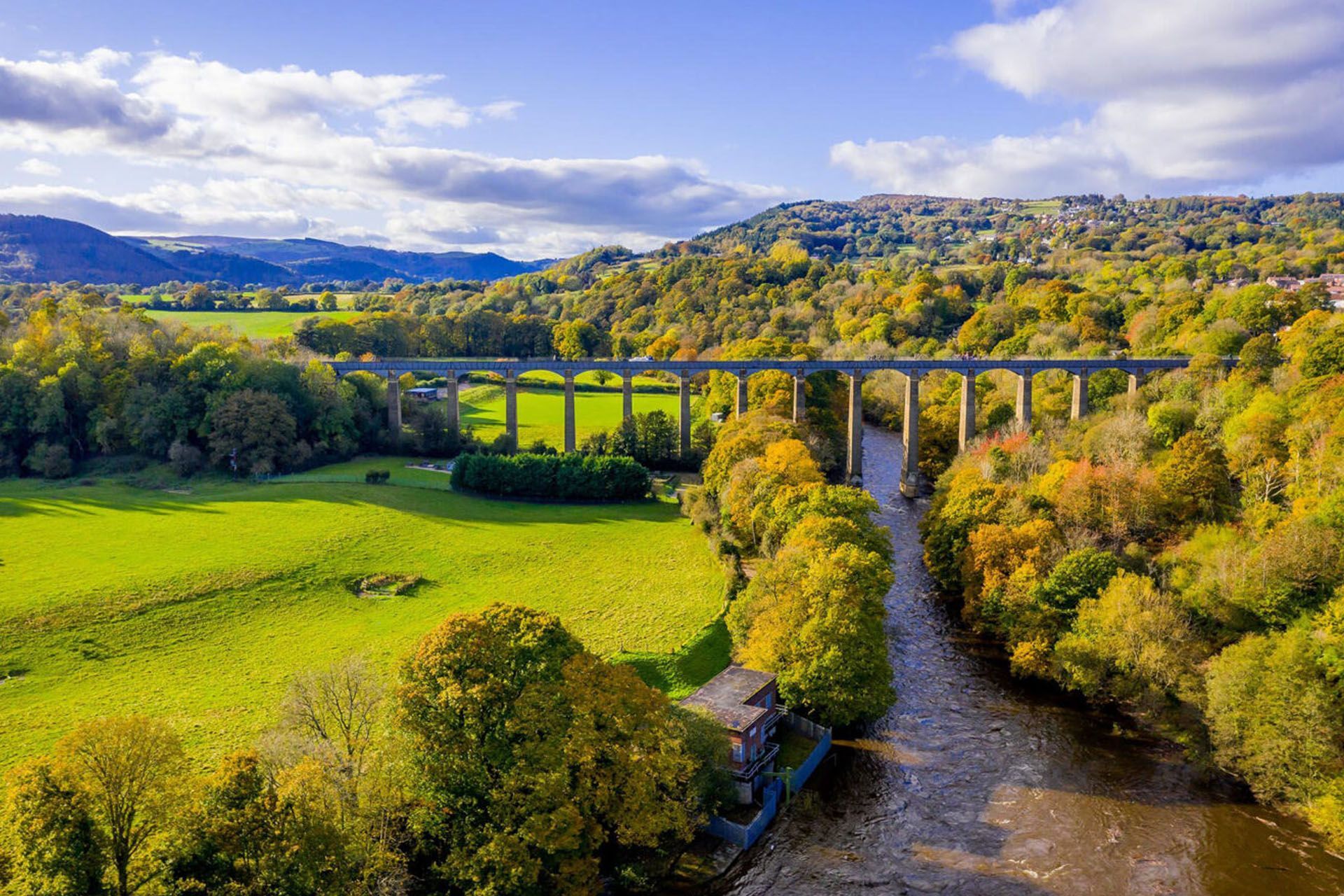 Sunlit river valley with a stone viaduct, green fields, trees, and hills under a cloudy sky