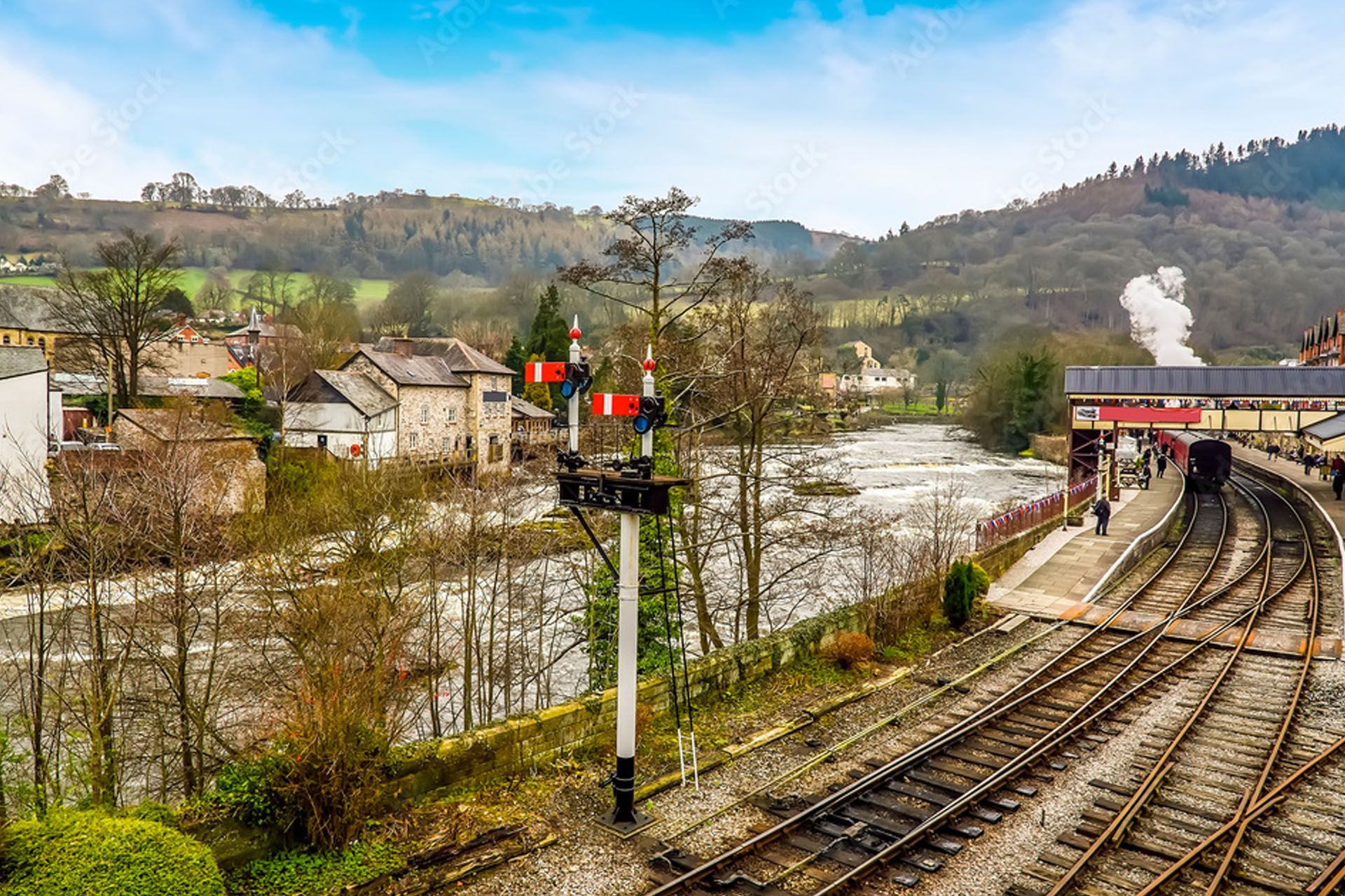 Steam train at a rural station beside tracks, with houses, trees, and hills in the background
