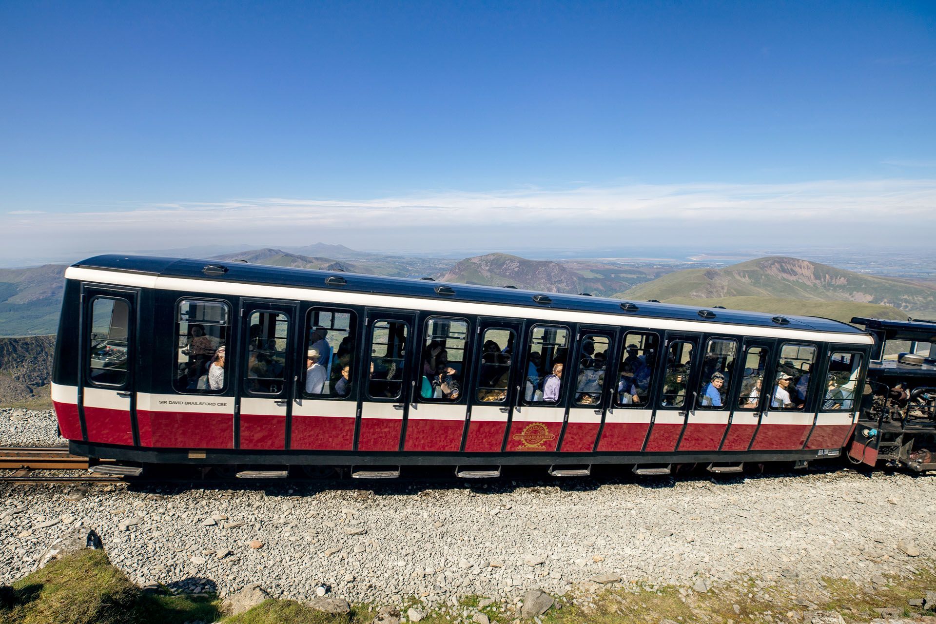 Mountain tram car with passengers, red and white, traveling along a rocky hillside under a clear blue sky